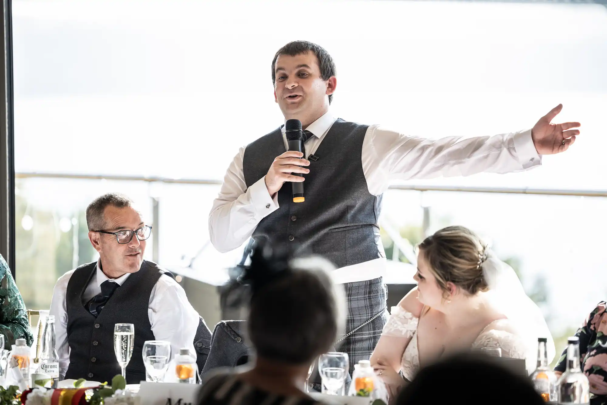 A man in a grey waistcoat and tie stands at a wedding reception, holding a microphone and gesturing with one arm. He speaks while seated guests, including a woman in a white dress and veil and an older man in spectacles, listen attentively at a decorated table.