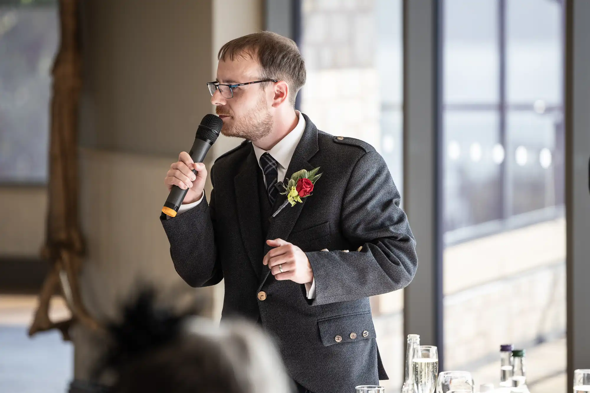 A man in a grey suit with a red rose buttonhole and striped tie speaks into a microphone at an indoor event. He stands beside a table with glassware, in front of large windows allowing bright natural light in.