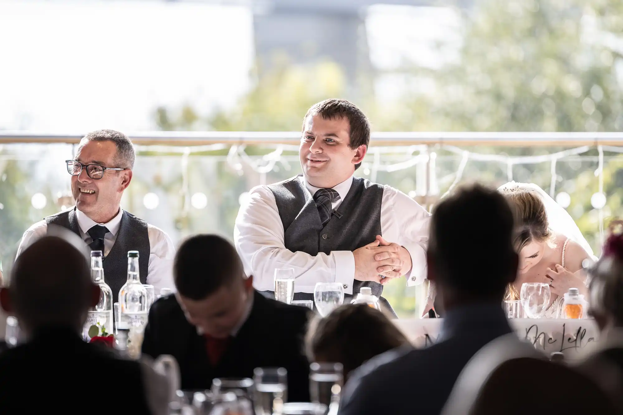 A smiling groom in a waistcoat and tie sits between an older man in glasses and a bride with her head down at a sunlit wedding reception table, decorated with fairy lights and glass bottles, with guests in the foreground.