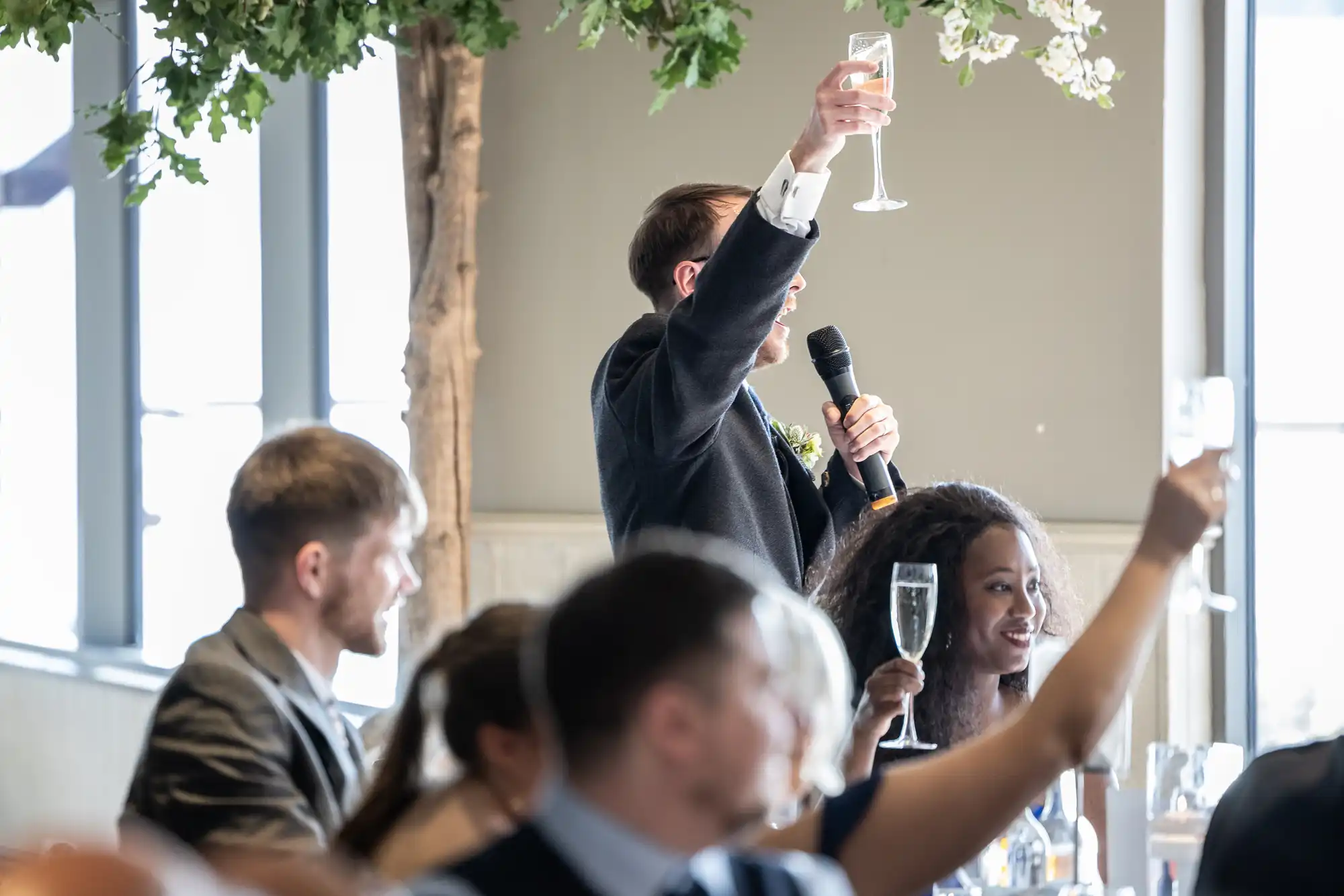 A man in a suit stands holding a microphone and raising a champagne glass in a toast at a wedding reception. Guests around him, seated at tables, also raise their glasses. Sunlight streams in through large windows and leafy decorations hang above.