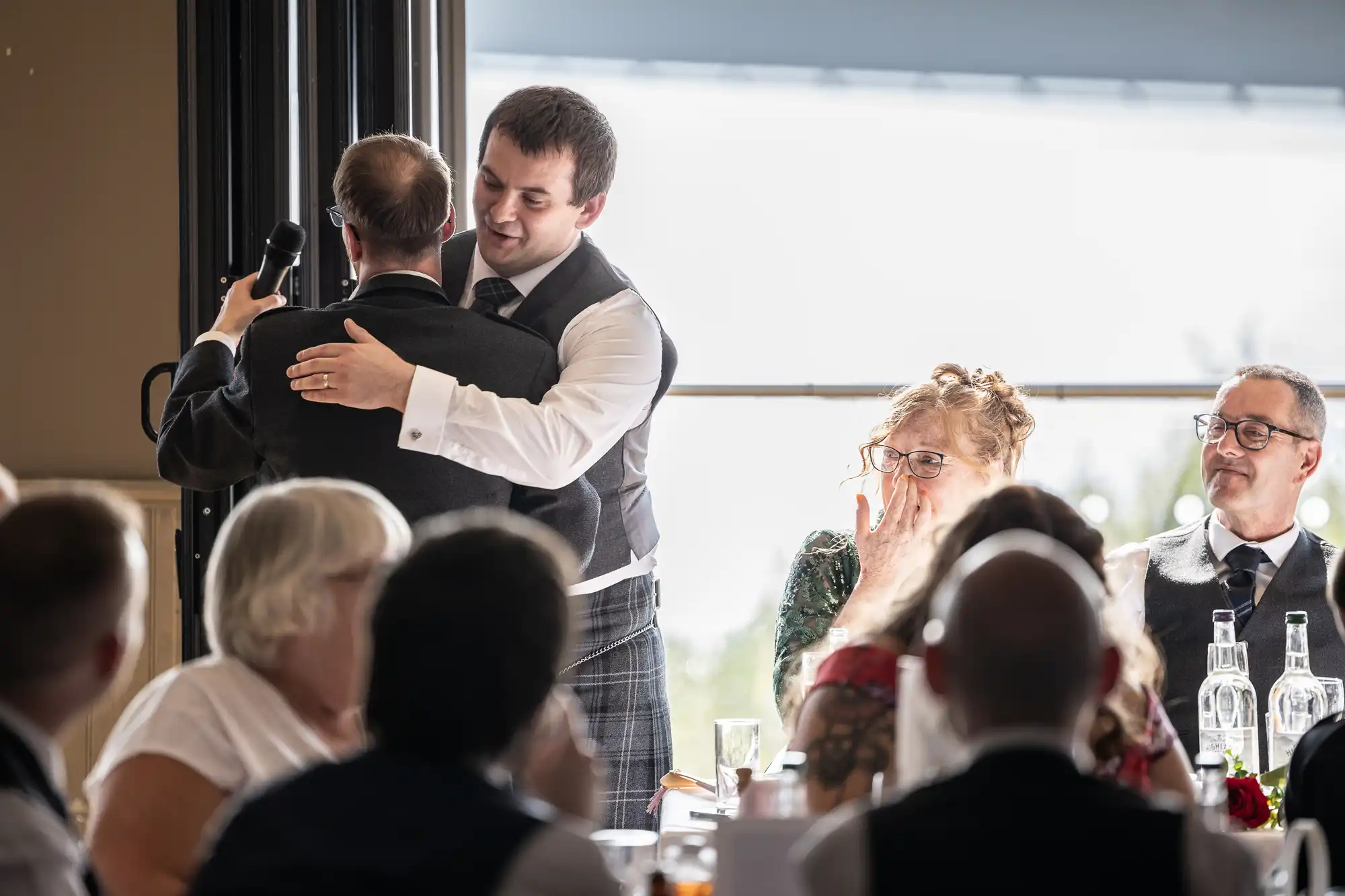 A man in a waistcoat hugs another man holding a microphone at a formal event, possibly a wedding. Seated guests watch, including a woman covering her mouth emotionally and a man in glasses smiling. Light streams in from a large window behind them.