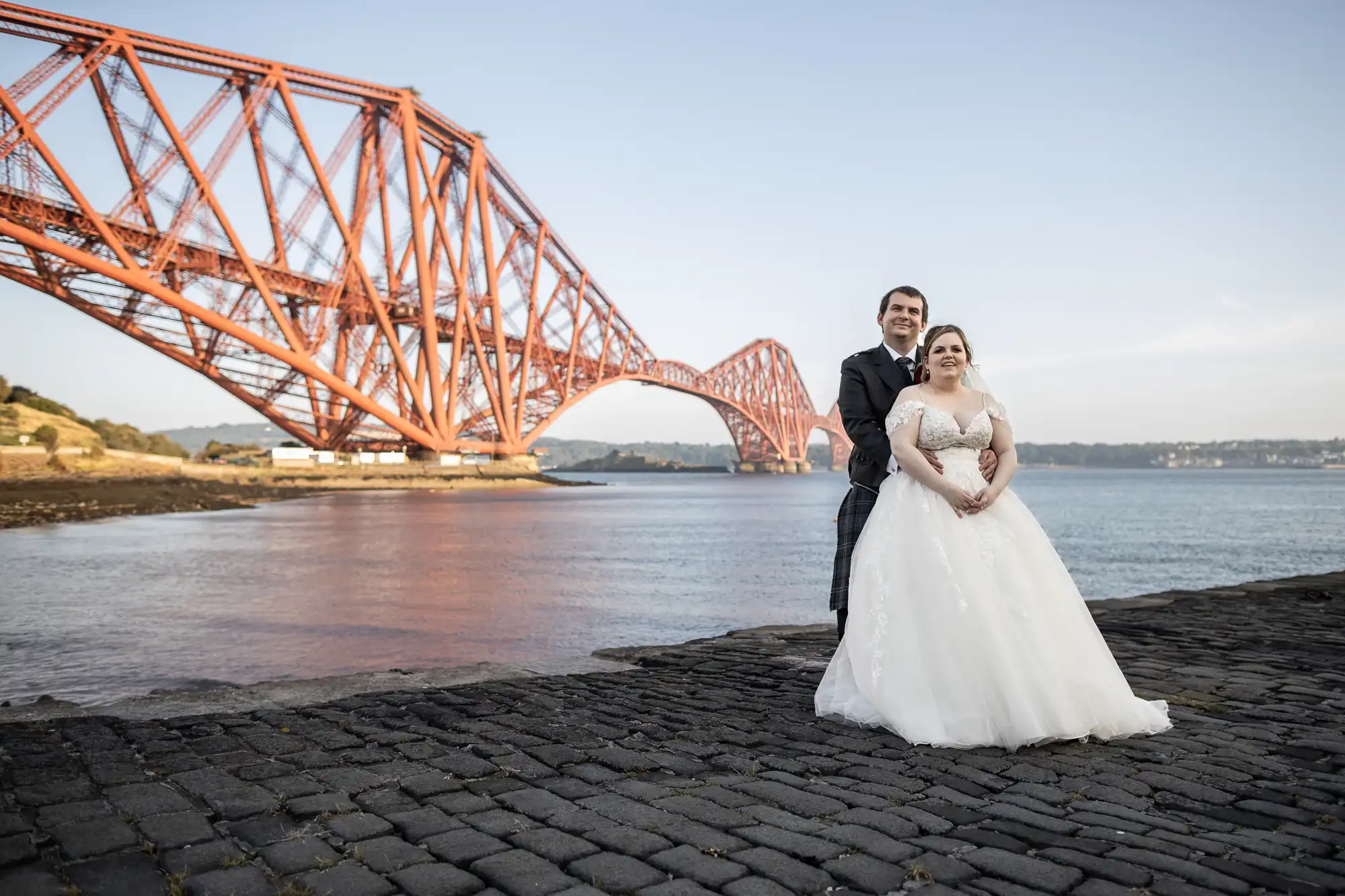 A bride in a white gown and a groom in a dark kilt stand on cobbles by the water, embracing and smiling, with the large red Forth Bridge and blue sky in the background.