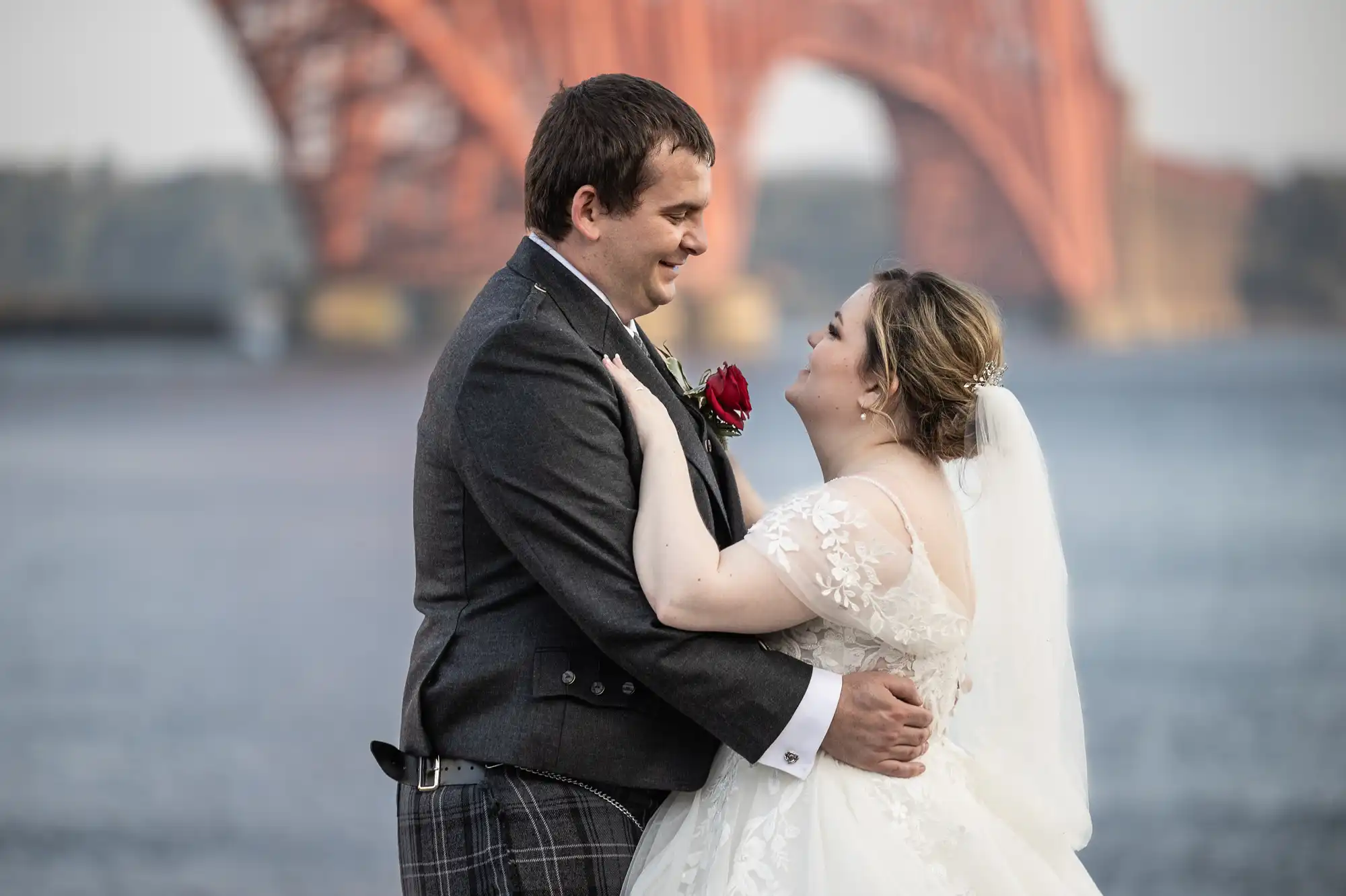 A bride and groom stand close, smiling at each other by the water. The bride wears a white dress and veil, and the groom is in a dark suit with checked trousers and a red rose buttonhole. An orange bridge arches in the blurry background.