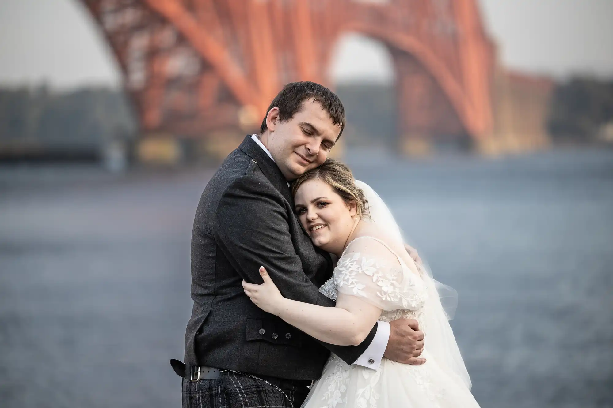 A newlywed couple embrace and smile outdoors near a river, with a large red-orange steel bridge in the background. The groom wears a dark suit with a kilt, and the bride wears a white dress and veil. The mood is joyful and loving.