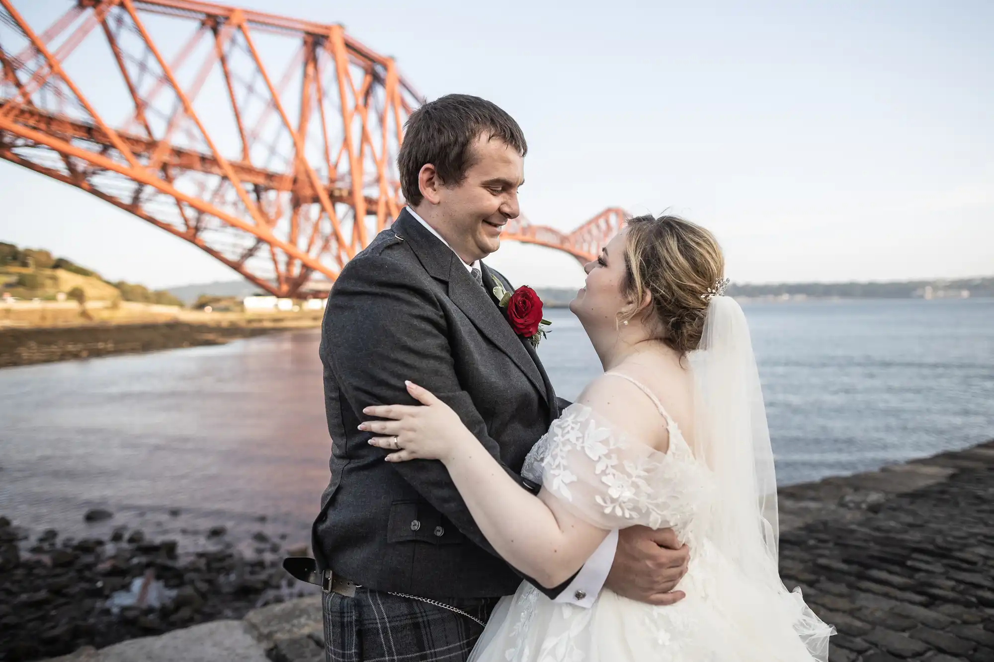 A bride and groom embrace and smile at each other near a waterfront, with the iconic red Forth Bridge in the background. The bride wears a white dress and veil; the groom is in a grey suit with a red rose buttonhole. It is a sunny day.