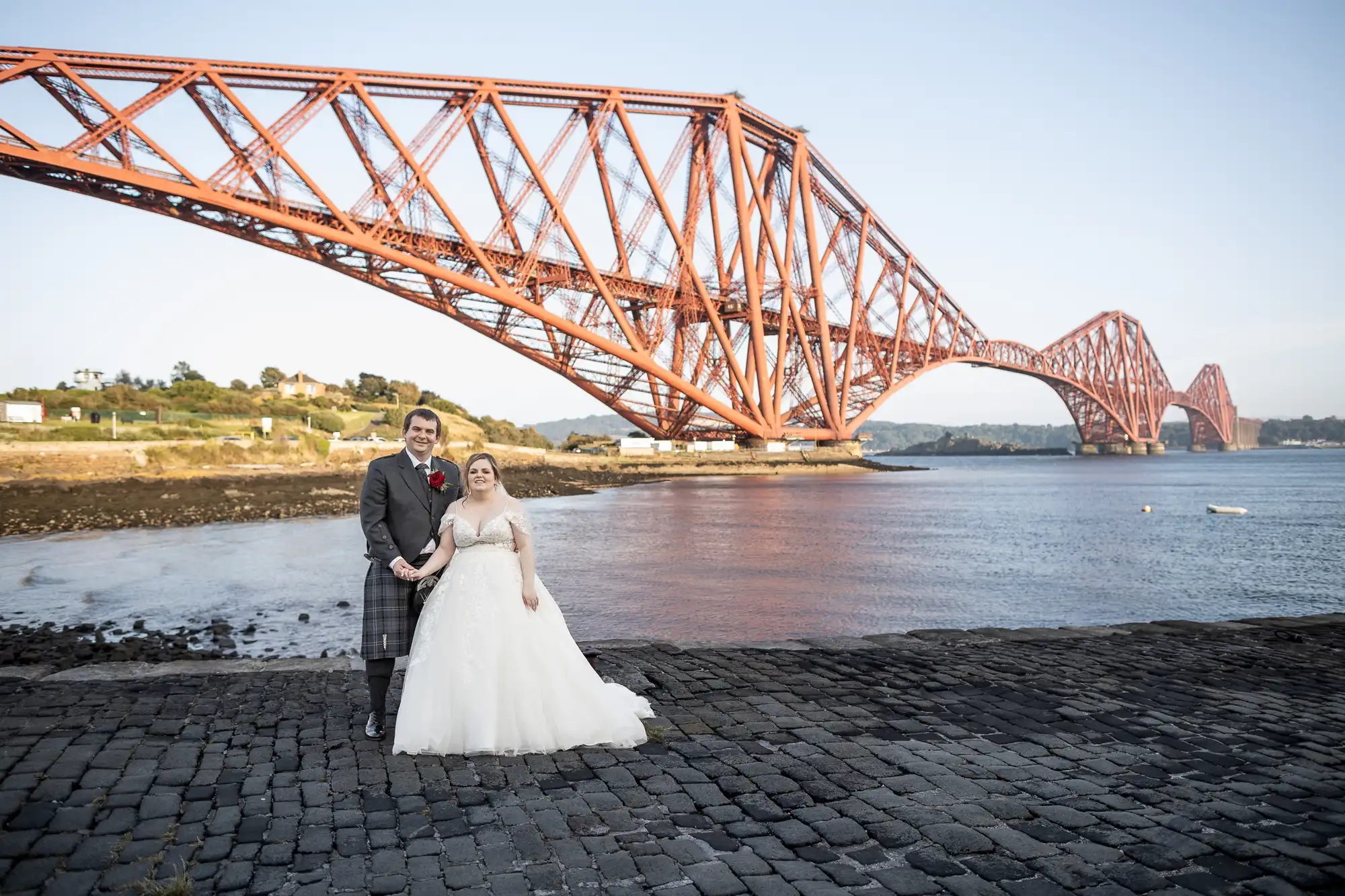 A bride in a white gown and a groom in a kilt pose together on a cobbled shore, smiling. Behind them is the iconic red Forth Bridge spanning across the water under a clear sky, with a hilly landscape in the background.