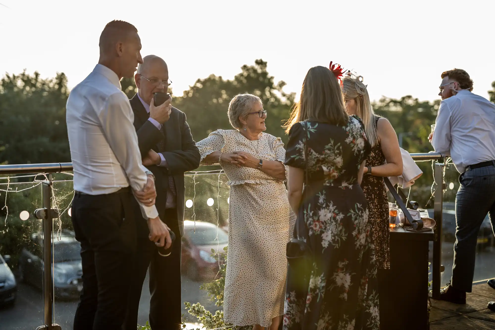 A group of six adults, dressed in semi-formal attire, stand outdoors in conversation at sunset. One woman in a white dress and glasses smiles whilst others, some holding drinks or mobiles, listen. Cars and trees are visible in the background.