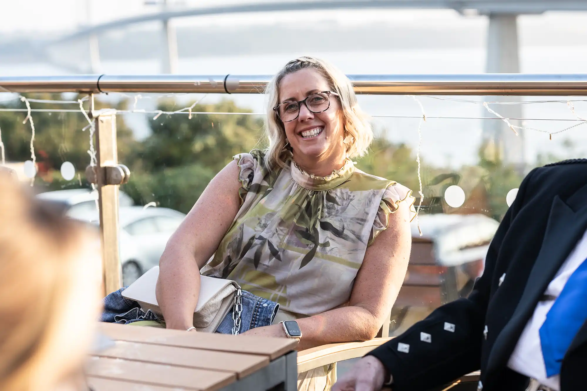 A smiling woman with glasses and wavy blonde hair sits outdoors on a patio, wearing a floral sleeveless top. She has a smartwatch on her wrist and a light-coloured bag on her lap. Decorative string lights and a bridge are visible in the background.