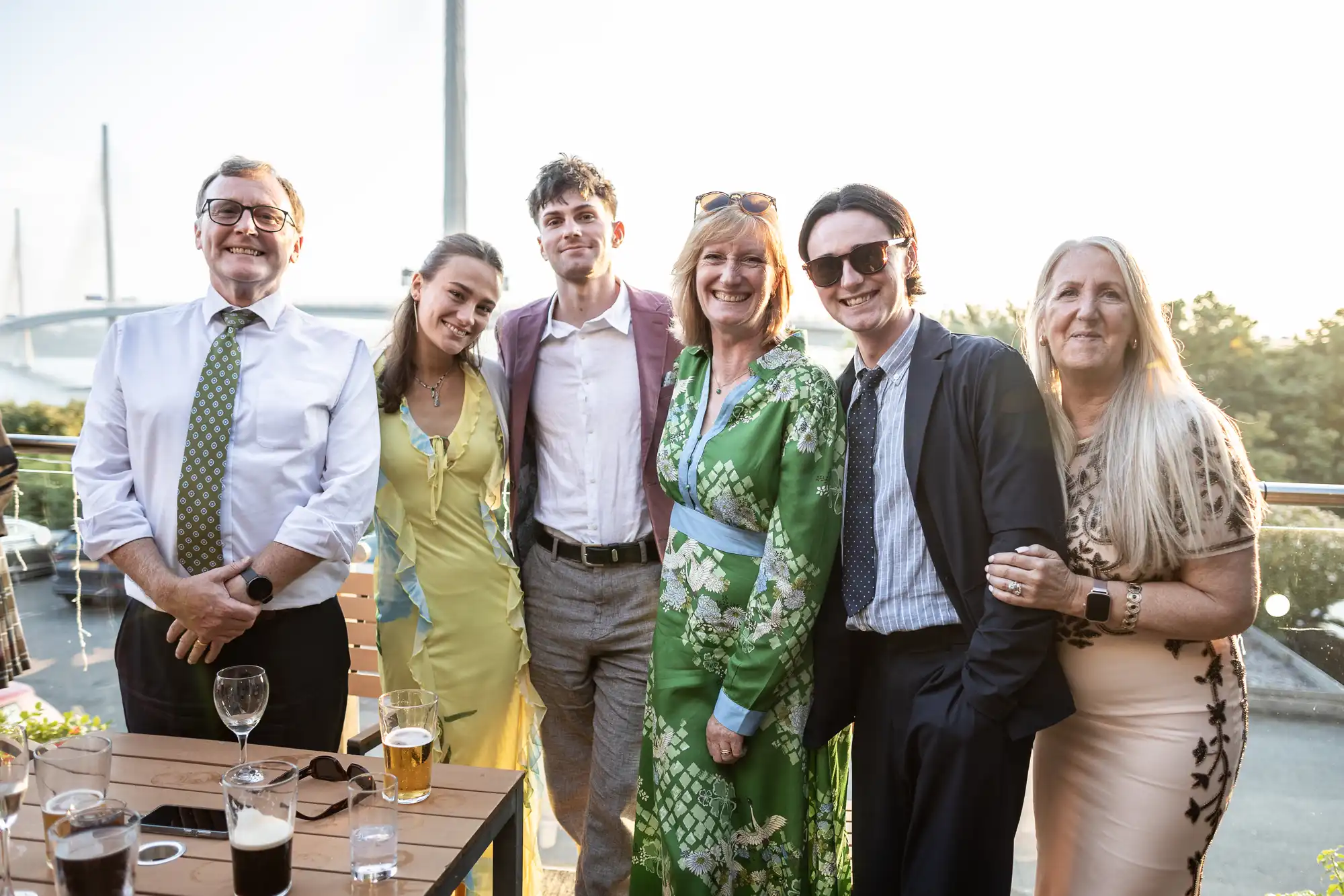 Six people stand closely together, smiling on a sunny outdoor patio. They wear semi-formal attire: dresses, suits, and ties in bright, stylish colours. Glasses of wine and beer sit on the wooden table in front of them. A bridge and trees are visible in the background.
