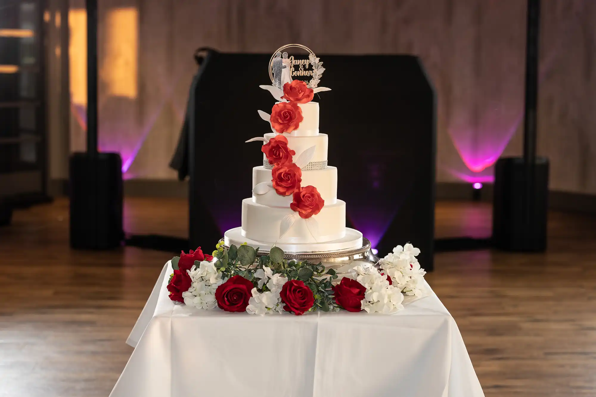 A three-tiered white wedding cake decorated with cascading red roses and a clear topper that says “Happily Ever After” sits on a round silver stand atop a white tablecloth. Red and white flowers surround the base; purple lights glow in the background.