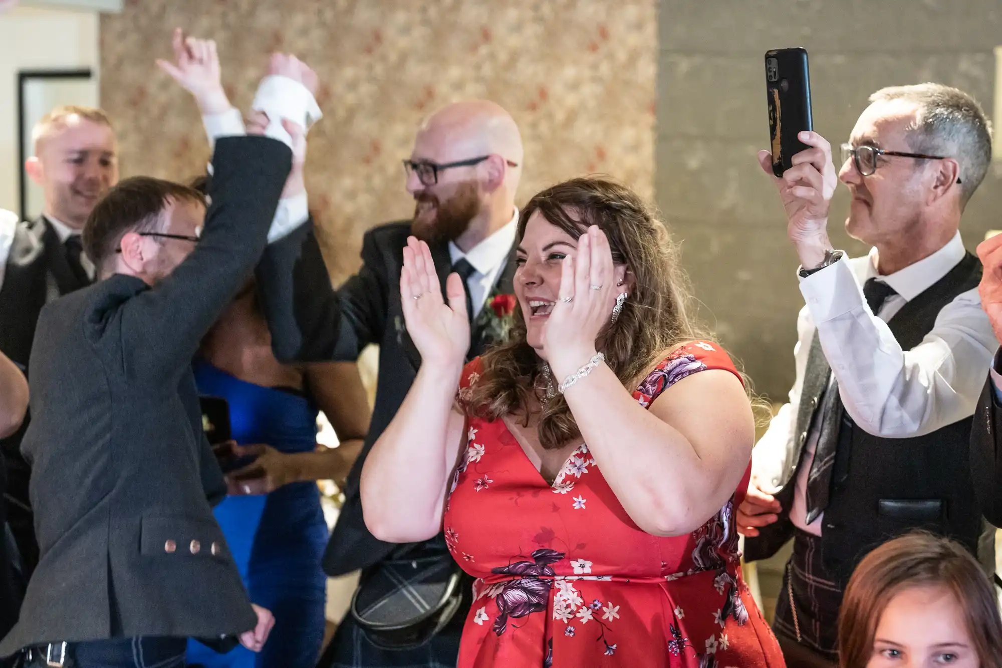 A joyful group of people celebrate indoors. A woman in a red floral dress claps her hands and smiles. Around her, men in suits and kilts cheer, one person raises their arms, and another takes a photo with a mobile. The scene is lively and festive.