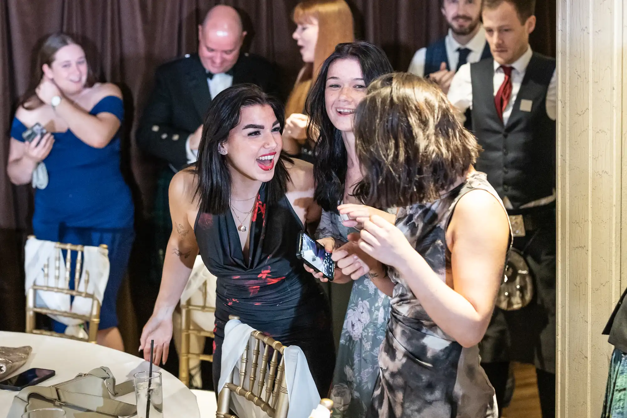 Three women in dresses stand by a table, smiling and laughing together at a lively indoor event. Other well-dressed guests mingle in the background, including men in suits and kilts. The atmosphere is festive and warm, with wooden chairs and soft lighting.