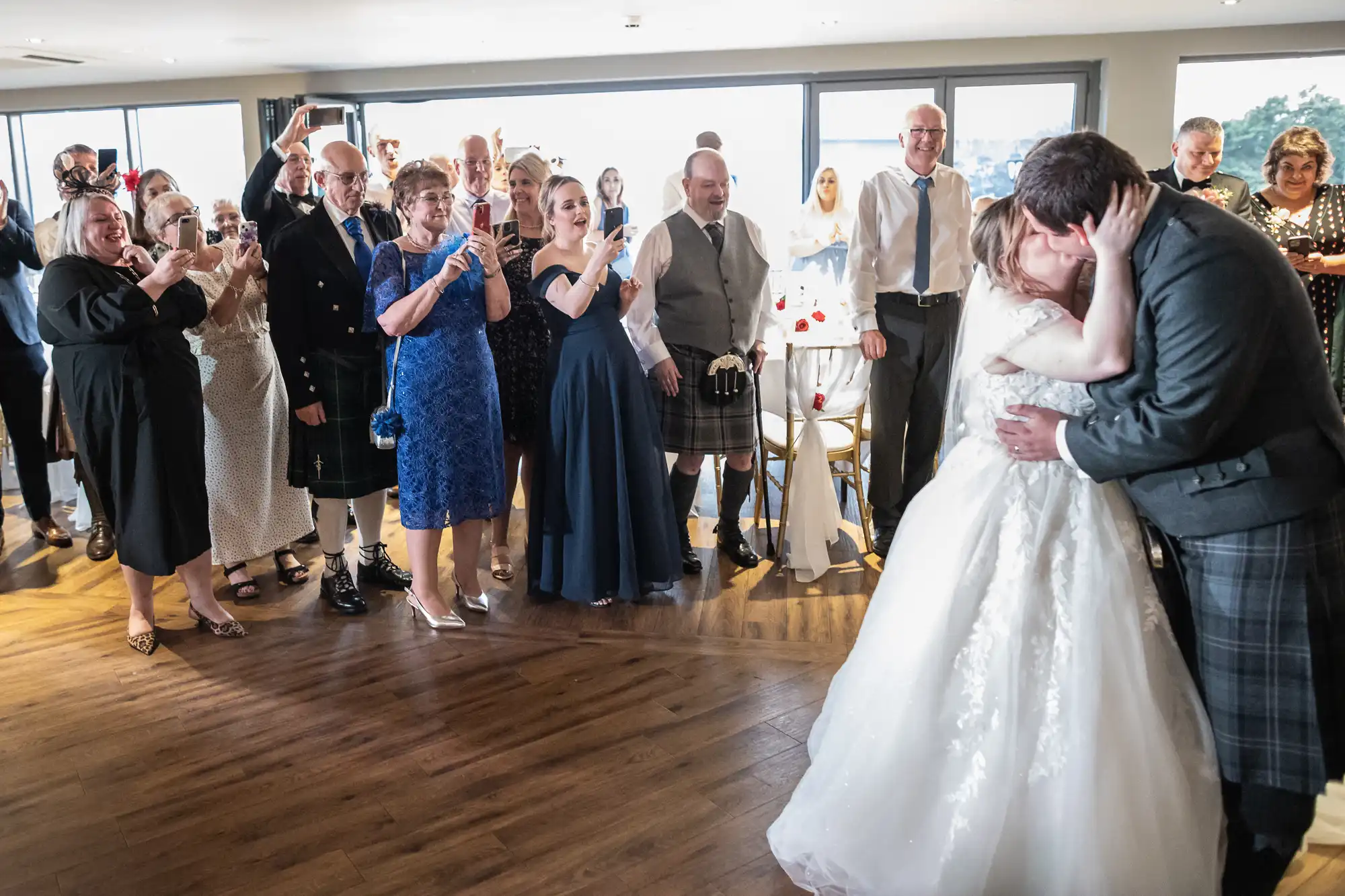 A bride and groom share a kiss on a dance floor in a bright room, surrounded by smiling and clapping guests dressed in formal attire. Some guests wear kilts, and there is a mix of blue and neutral-coloured clothing. Sunlight streams in from large windows behind them.