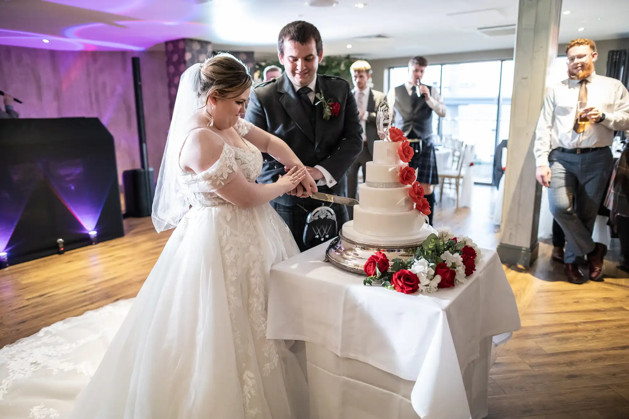 A bride and groom, both smiling, cut a white tiered wedding cake decorated with red and white roses. The bride wears a lace gown and glasses; the groom is in a dark suit. Guests watch and celebrate in a bright, elegant venue.