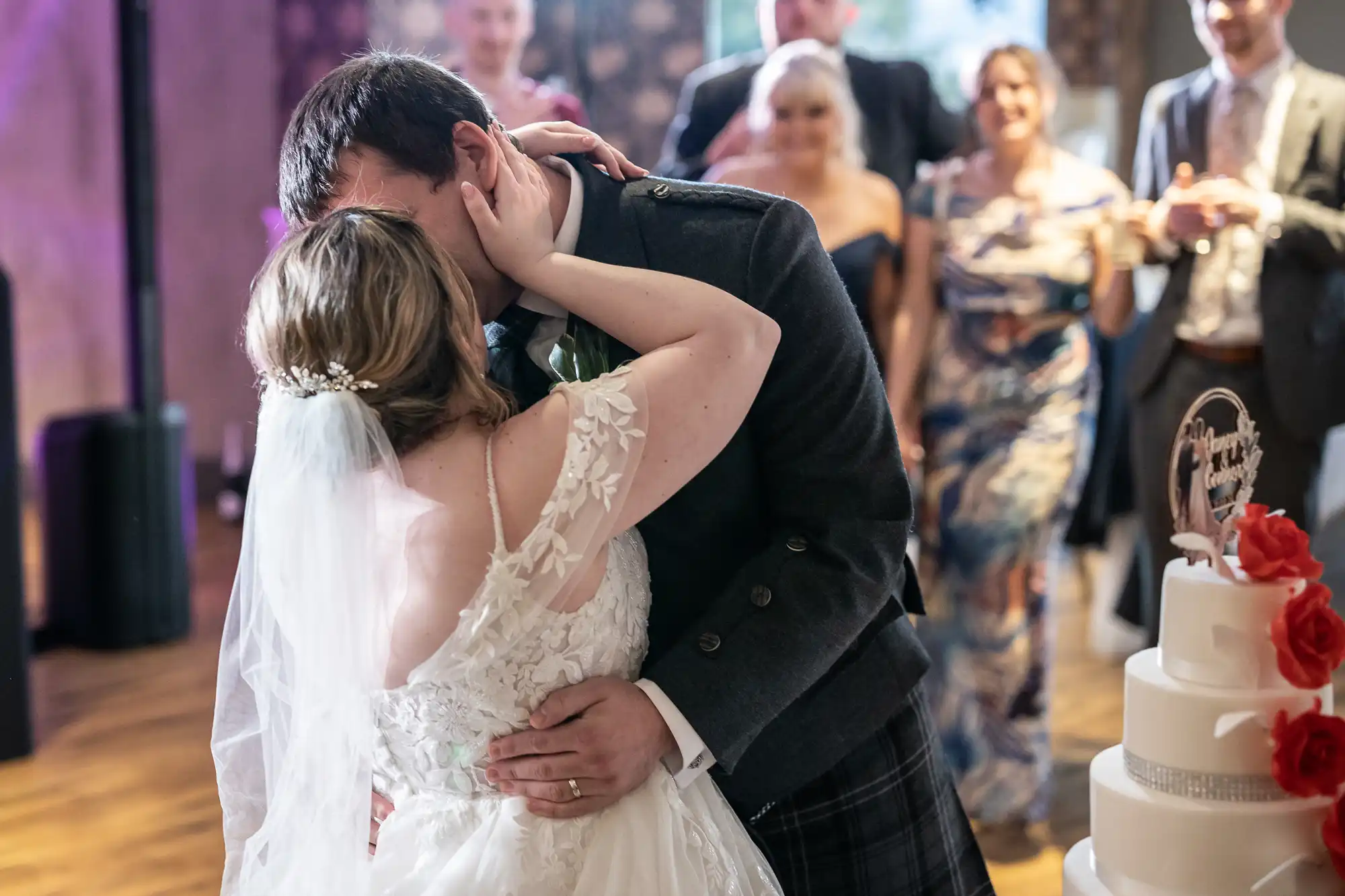A bride and groom share a kiss at their wedding, with the bride wearing a white lace dress and veil. The groom is in a dark suit. Guests in formal attire watch and smile in the background. A tiered wedding cake with red flowers is visible in the foreground.