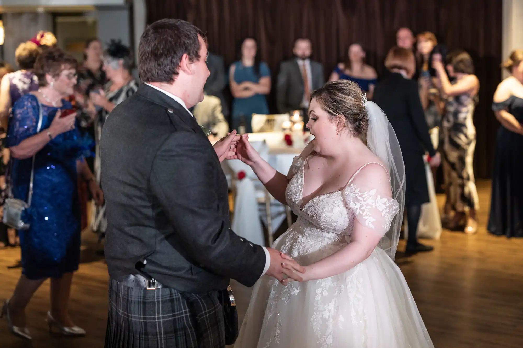 A bride and groom dance together at their wedding reception. The bride wears a white, off-the-shoulder gown with lace details and a veil. The groom wears a dark suit with a kilt. Guests in formal attire watch and celebrate in the softly lit background.