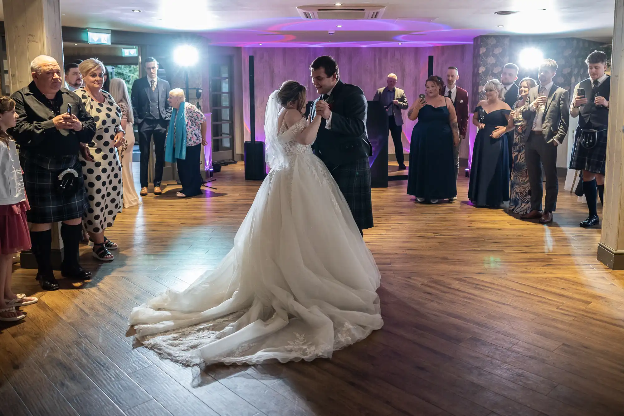 A bride and groom share their first dance in the centre of a wooden-floored room, surrounded by guests in formal attire, some wearing kilts. The bride wears a long white gown, and soft purple lighting illuminates the celebratory scene.