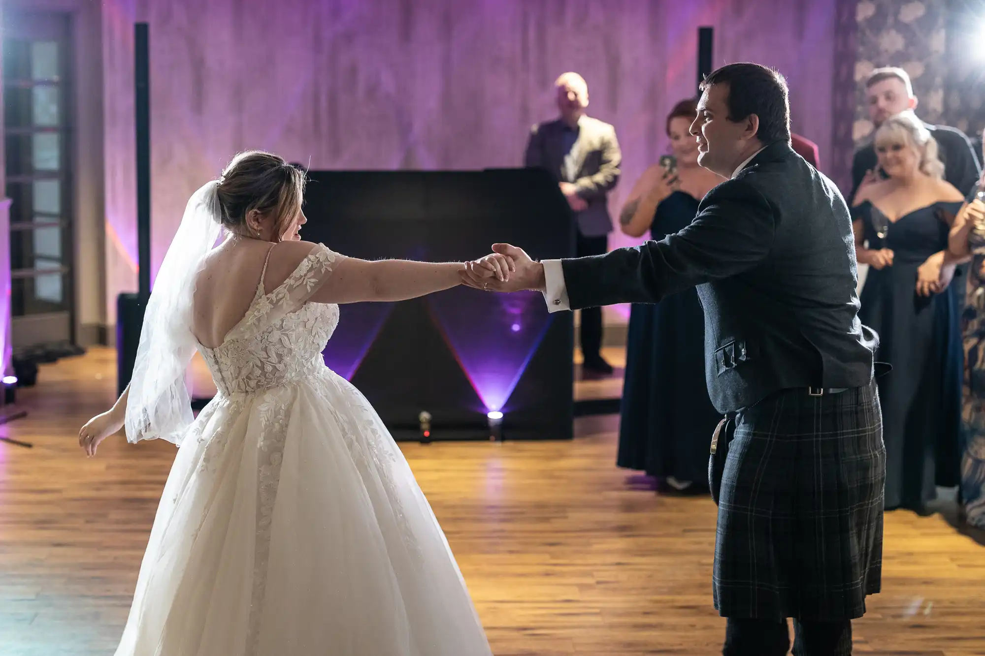 A bride in a white dress with a veil and a groom in a dark jacket and kilt hold hands while dancing. Guests in formal attire watch them on a wooden floor in a softly lit reception hall with purple lighting.