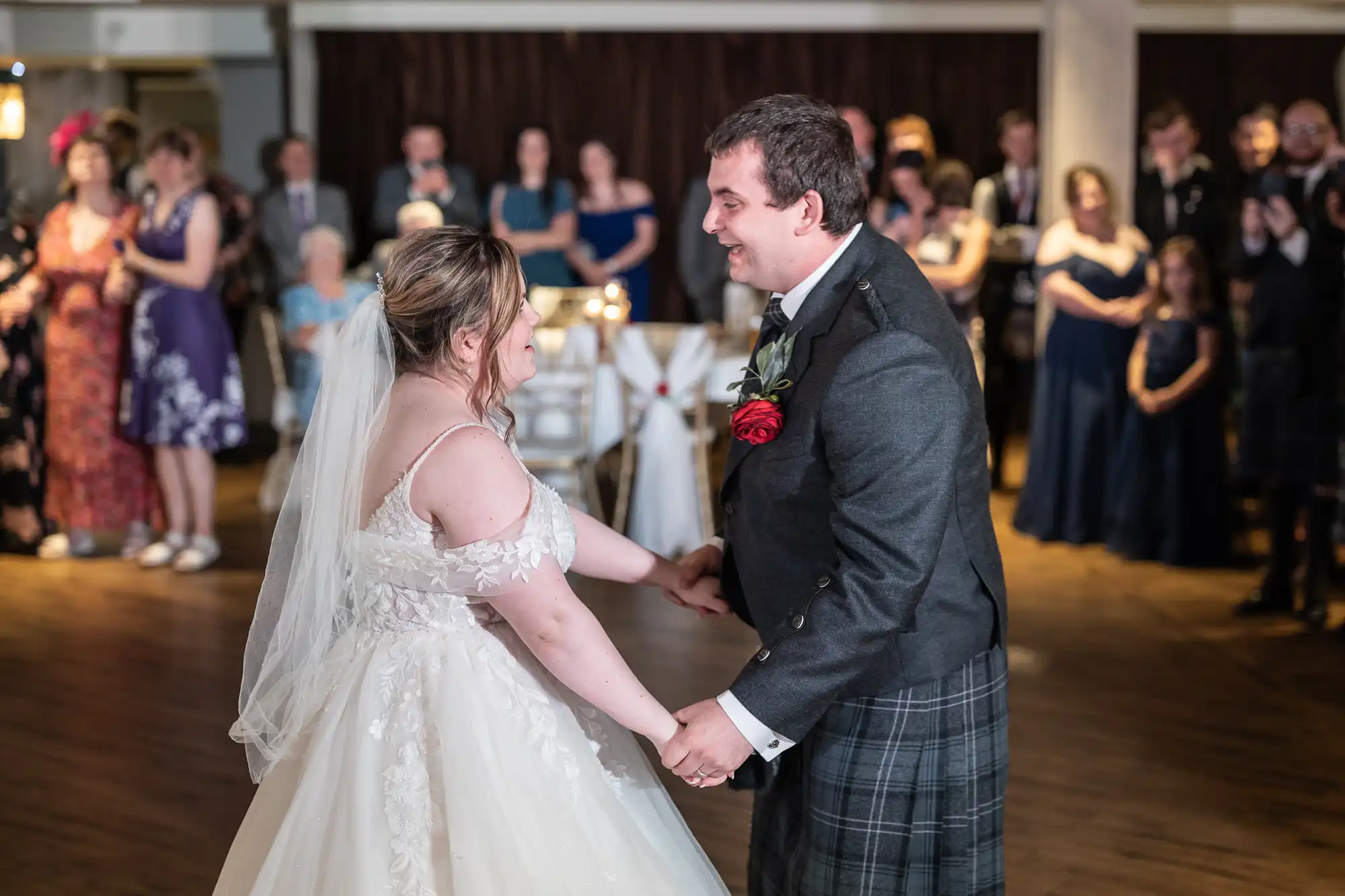 A bride in a white gown and veil dances, smiling, with a groom in a suit and kilt. They hold hands on a wooden floor, with guests in formal attire watching and smiling in the softly lit background.