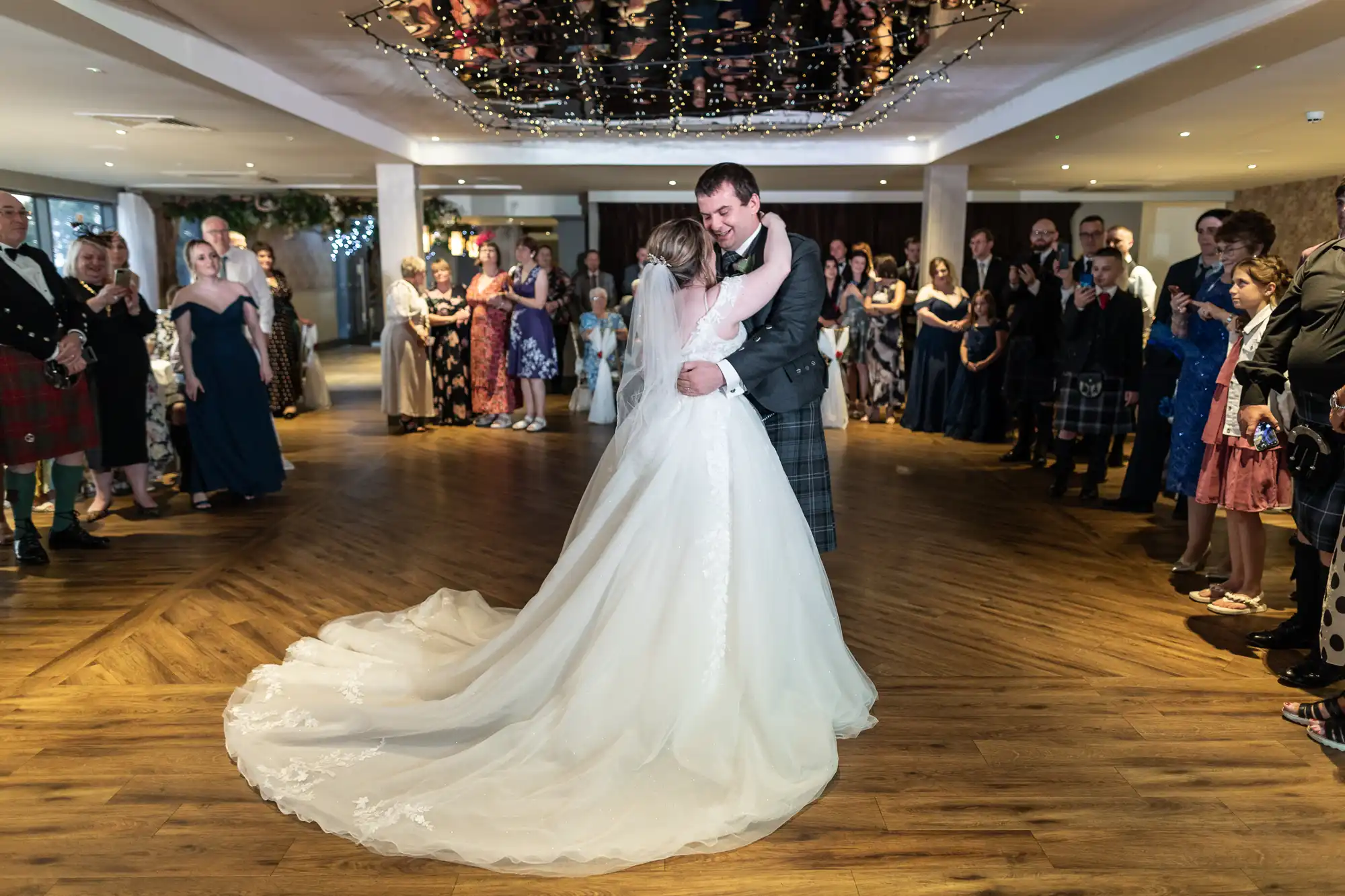 A bride and groom share their first dance in a warmly lit venue, surrounded by guests in formal attire. The bride wears a long white gown with a train, while the groom wears a suit with a tartan kilt. Fairy lights hang above, and guests watch, smiling.