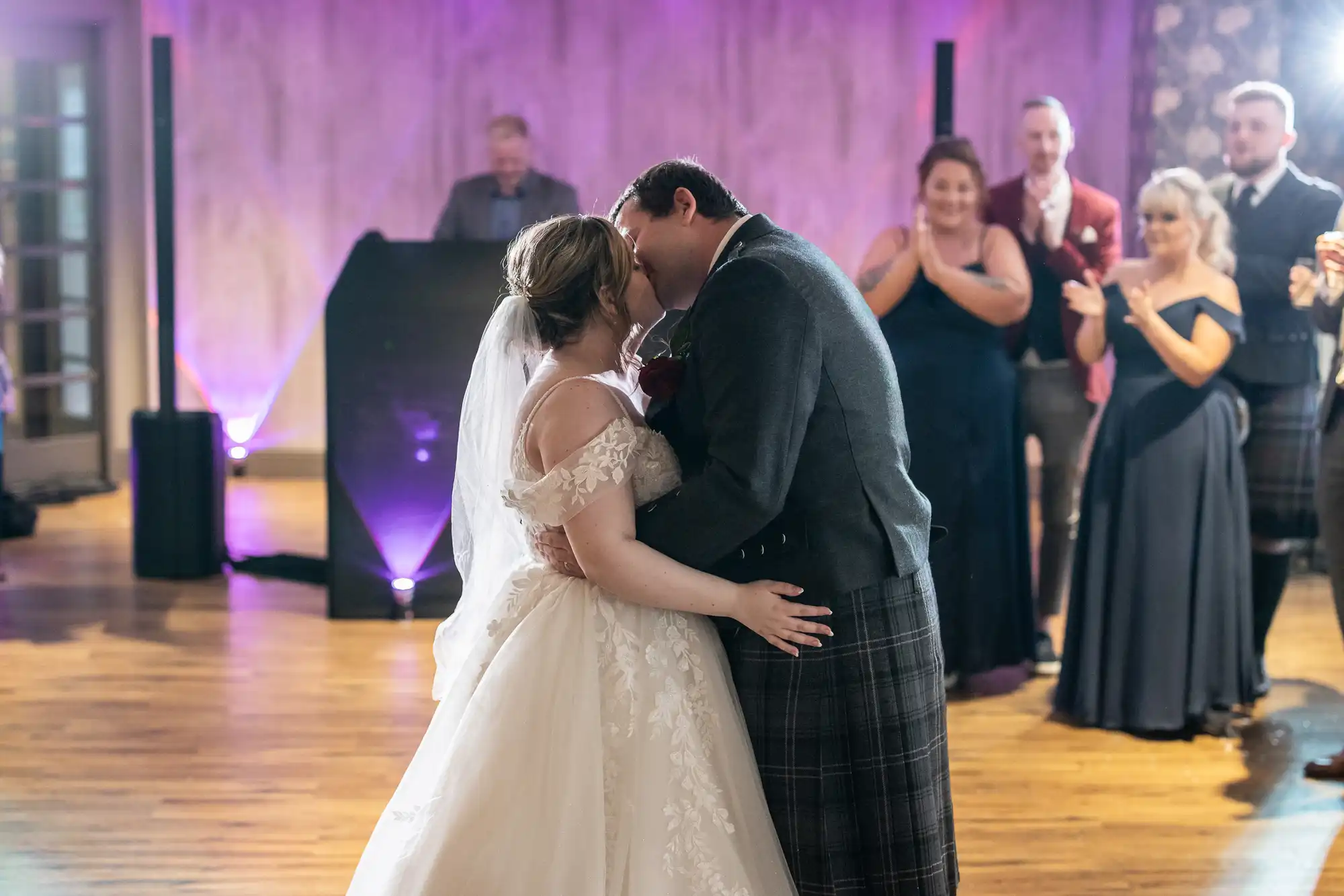 A bride and groom share a kiss on a dance floor during their wedding, surrounded by applauding guests in formal attire. The bride wears an off-shoulder white gown and veil, whilst the groom wears a dark jacket and tartan kilt. Soft purple lighting fills the room.