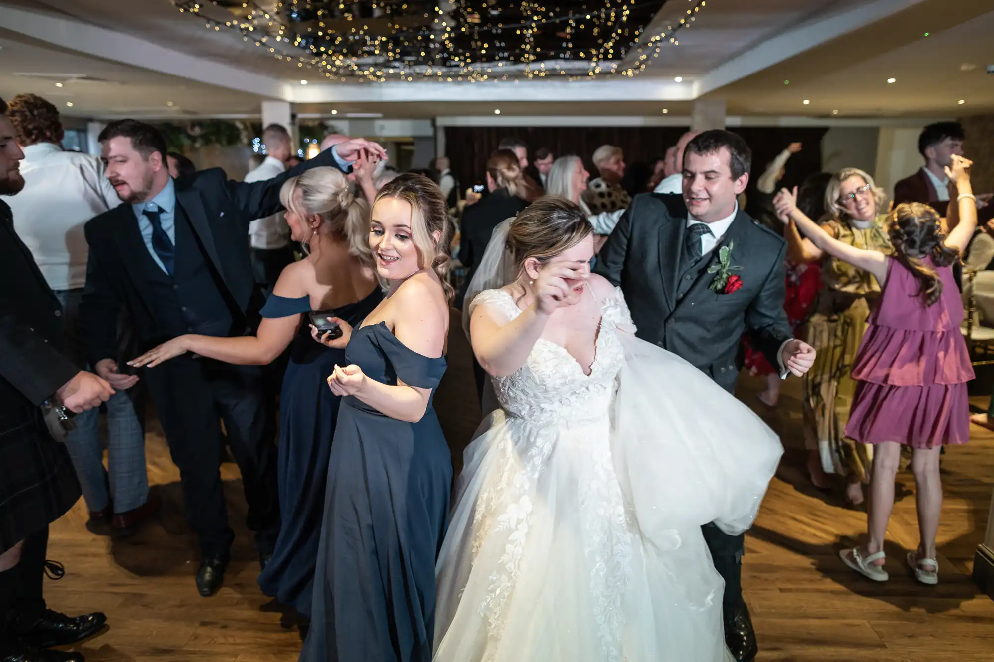 A joyful wedding reception scene with guests dancing. The bride in a white gown and a bridesmaid in a navy dress are at the centre, smiling and pointing as they dance. Other guests in formal attire dance and mingle under warm lights and hanging fairy lights.