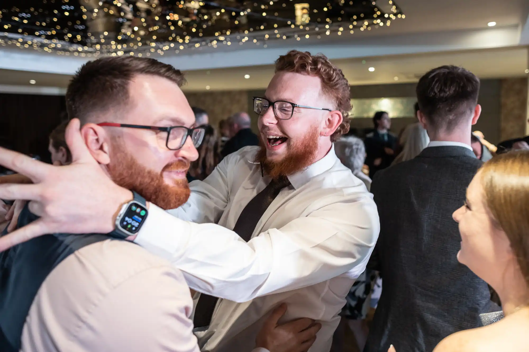 Two men with beards and glasses joyfully greet each other at a lively indoor party under twinkling fairy lights. One man, wearing a smartwatch and dress shirt, embraces the other. People in formal wear mingle and dance in the background.