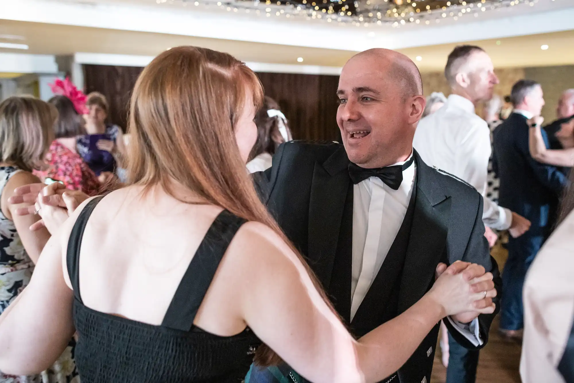 A man in a black dinner jacket and bow tie happily dances with a woman in a sleeveless black dress at a lively indoor event. Other guests chat and dance in the background under fairy lights. The atmosphere appears festive and joyful.