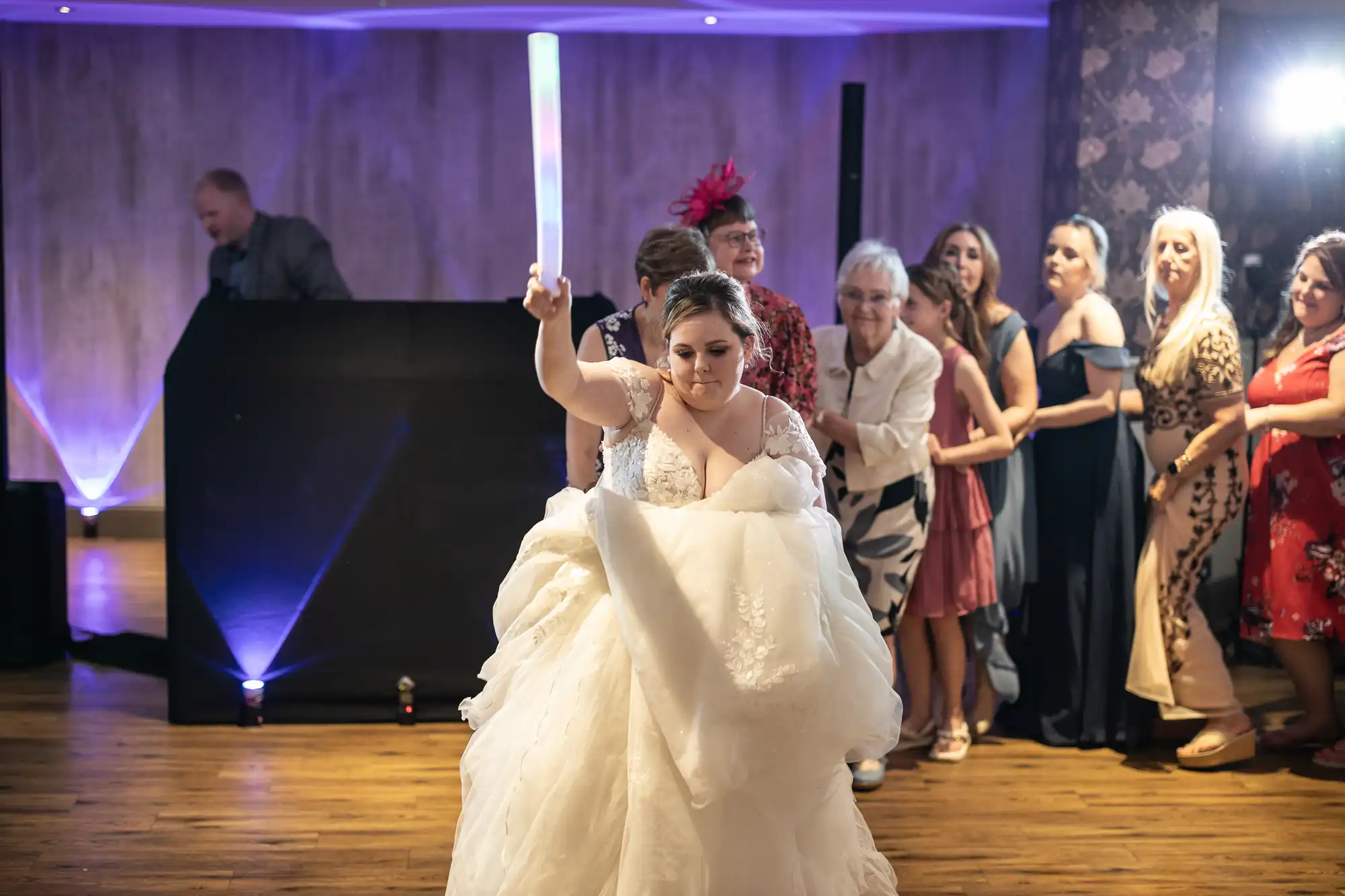 A bride in a white dress holds a glowing baton aloft on a dance floor, facing the camera. Behind her, women in elegant dresses smile and watch. A DJ stands at a booth in a softly lit room with purple uplighting on the walls.