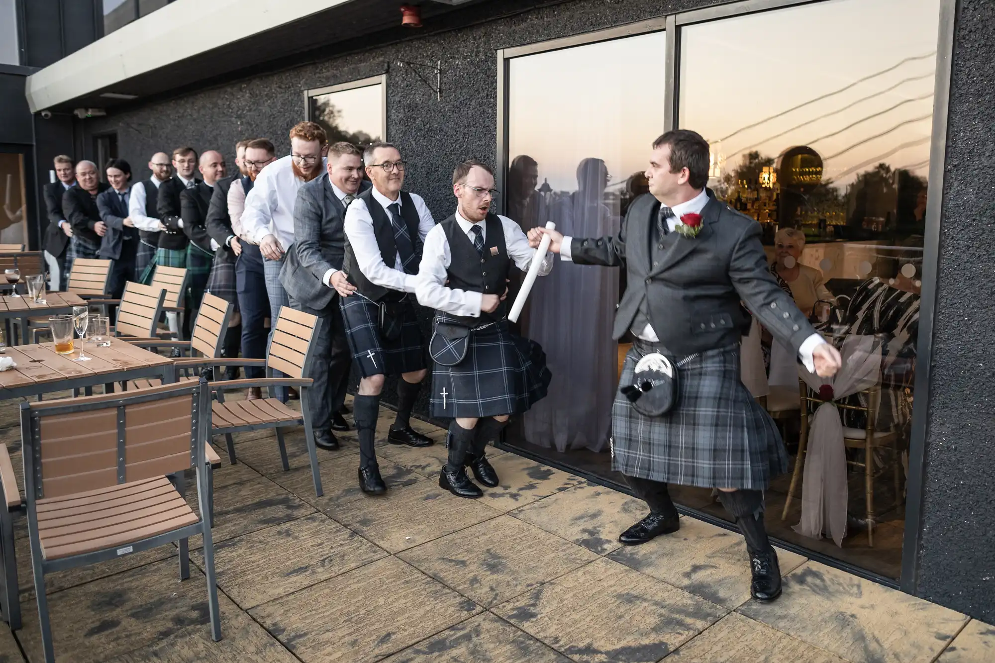 A group of men in kilts form a conga line on an outdoor terrace. The leader, holding a scroll and wearing a buttonhole, guides them. The men are smiling and laughing, reflected in the large windows. Wooden chairs and tables line the terrace.