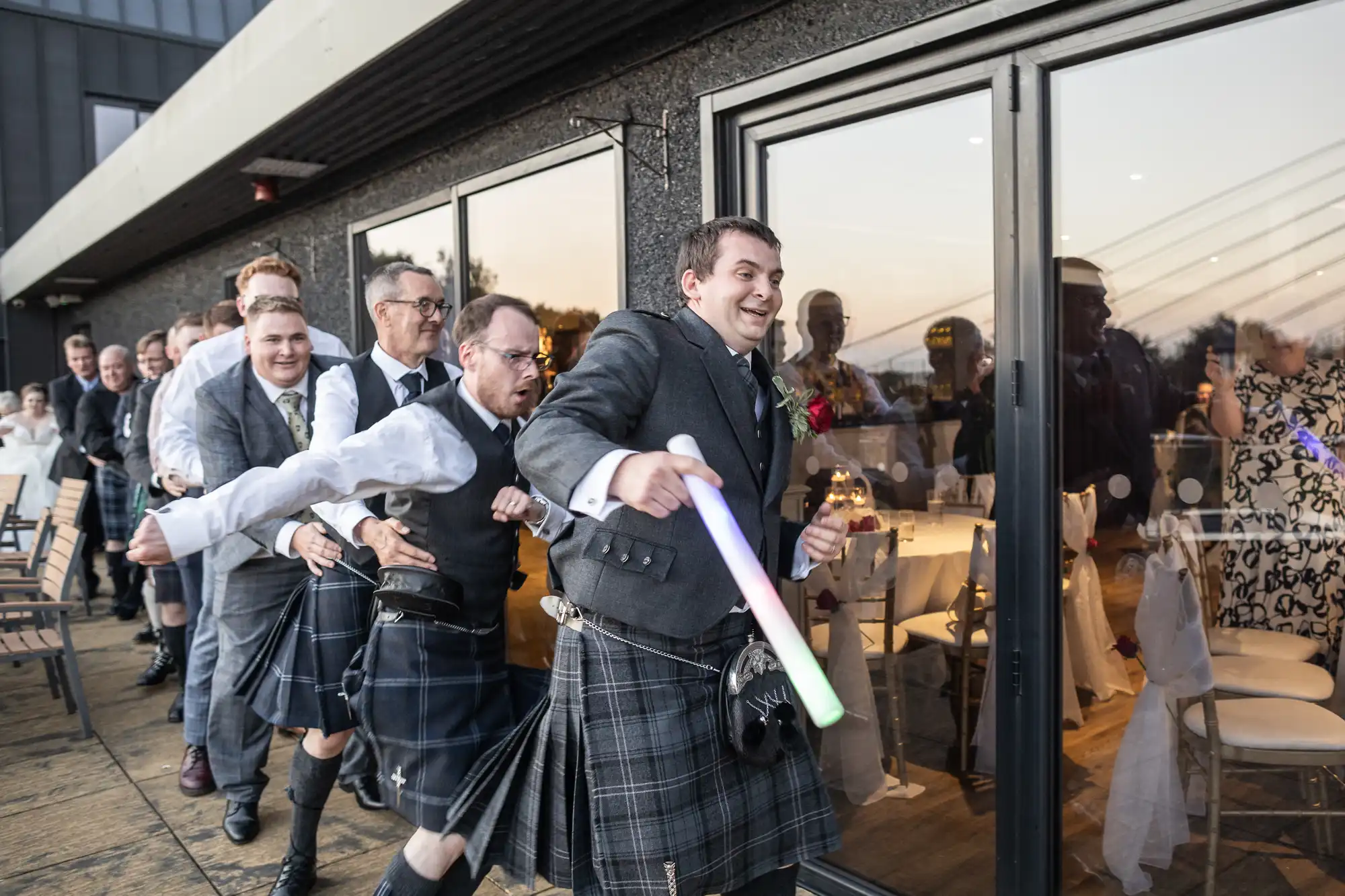 A group of men wearing kilts form a conga line outside a building with large glass doors. The leader smiles and holds a light-up baton. People inside and outside watch and laugh. The scene appears festive, likely at a wedding reception.