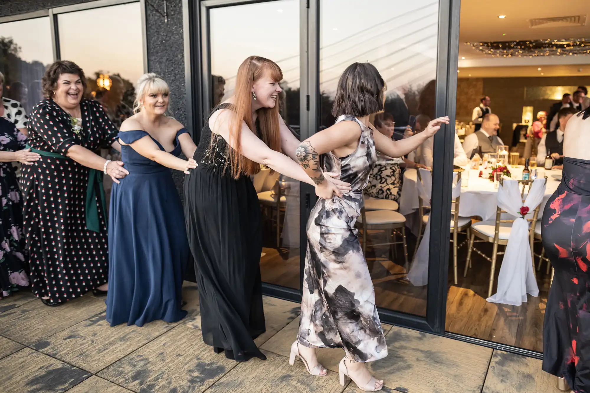A group of women in colourful dresses form a conga line, laughing as they dance outside glass doors of a banquet hall, where guests sit at round tables inside. The setting appears festive, with warm lighting and party decorations visible indoors.