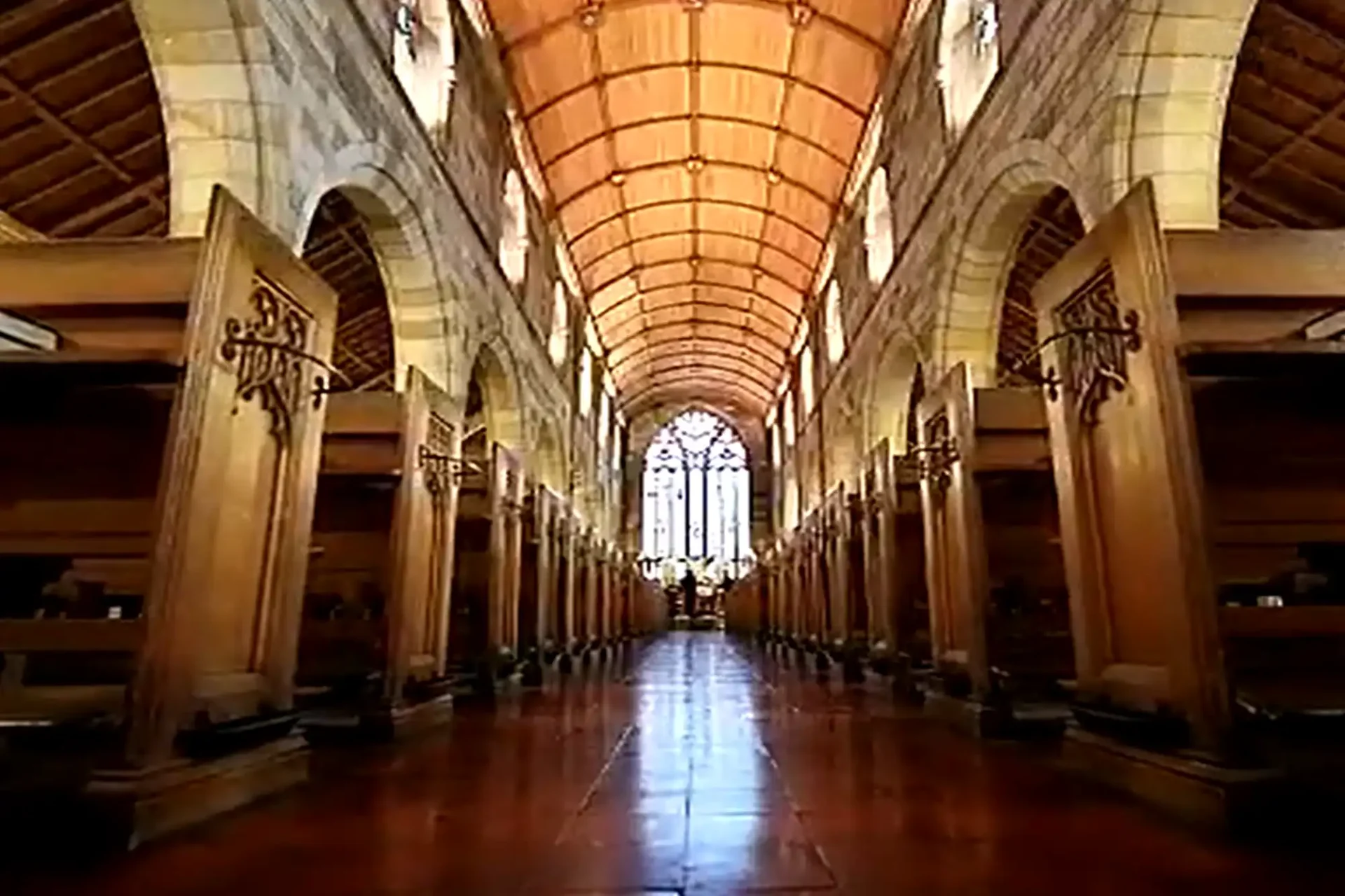 A wide view of a church interior features symmetrical wooden pews, tall stone arches, and a vaulted wooden ceiling. Sunlight streams through a large stained glass window at the far end, illuminating the polished tiled floor and ornate architectural details.