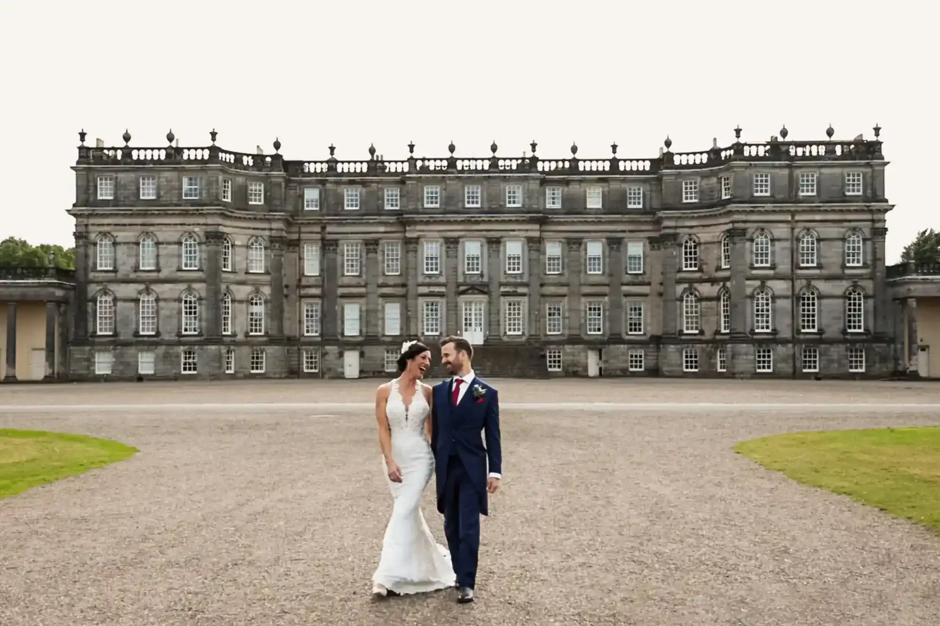 A bride and groom walk hand in hand in front of a large, historic stone building with many windows and decorative architecture.