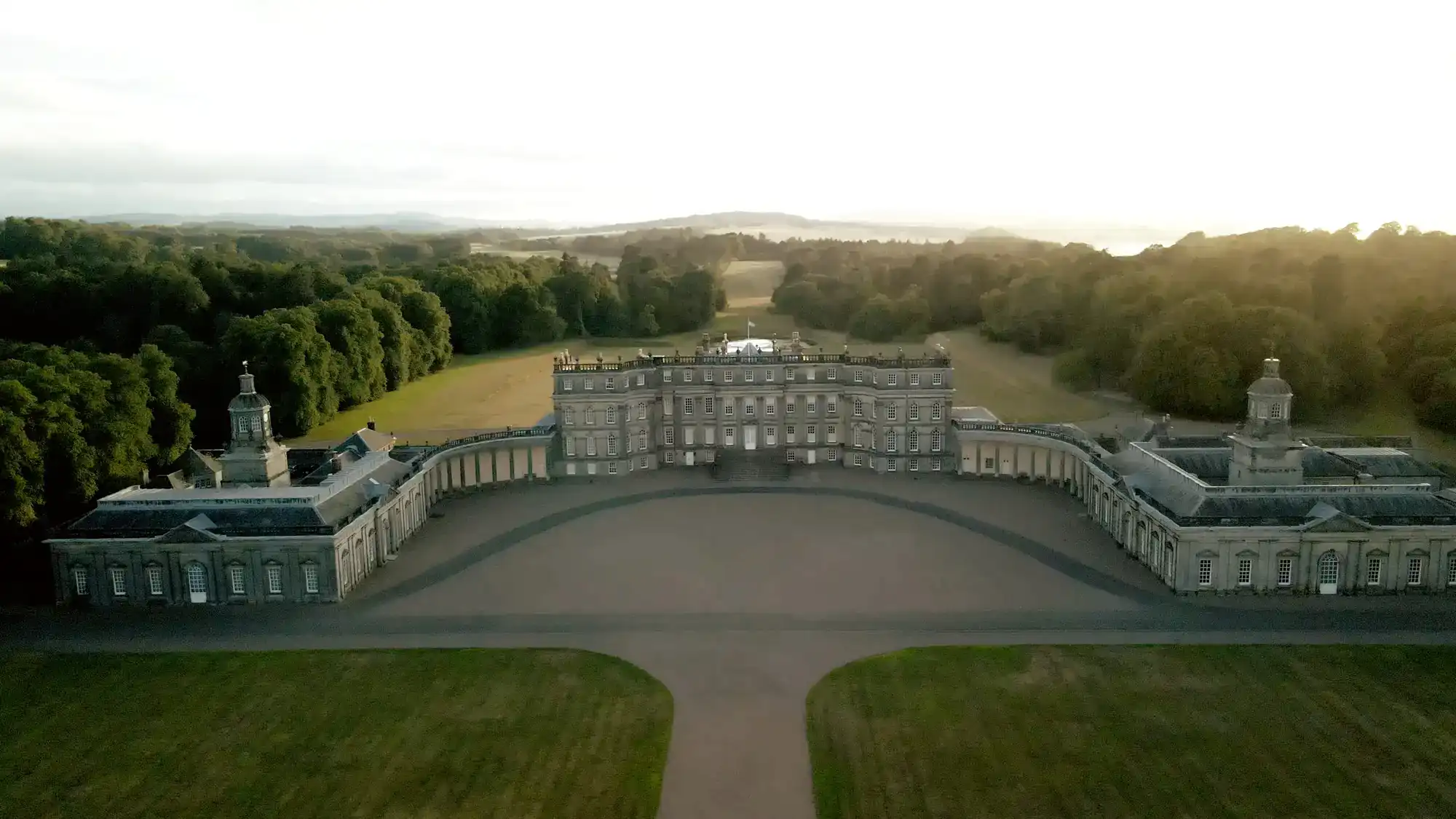 Aerial view of a grand, symmetrical historic mansion with two wings curving outward, set amid expansive green lawns and surrounded by dense trees under a pale, hazy sky at sunrise or sunset. Rolling hills are visible in the distant background. A drone photo created by award-winning Hopetoun House wedding videographer.