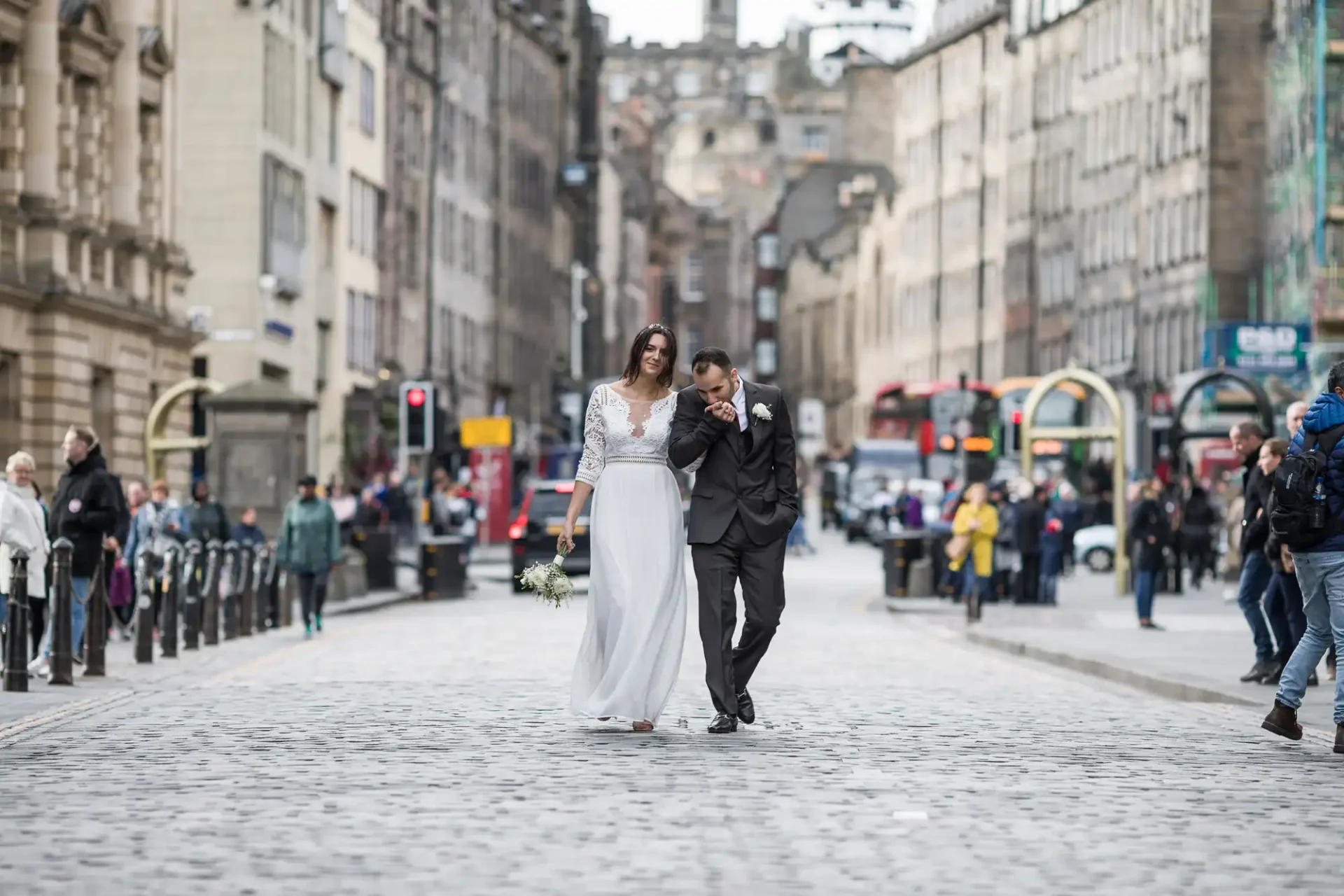 A couple walks arm in arm down a cobblestone street, surrounded by city buildings and pedestrians. The bride is in a white dress holding flowers, and the groom is in a black suit.