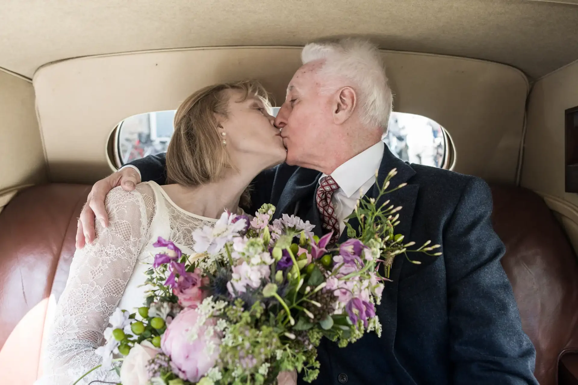 An older couple kisses in the backseat of a car, the woman holding a bouquet of flowers.