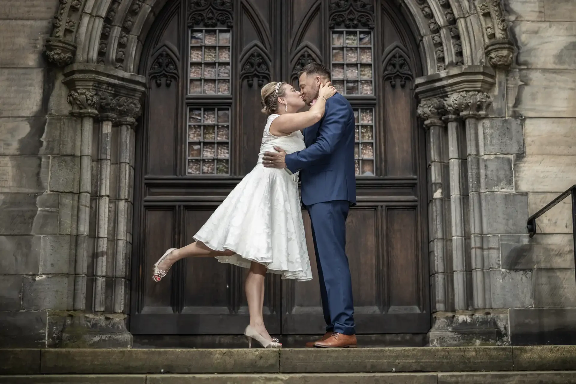 A couple embraces and kisses on the steps of a gothic-style building, with the woman in a white dress and the man in a blue suit.