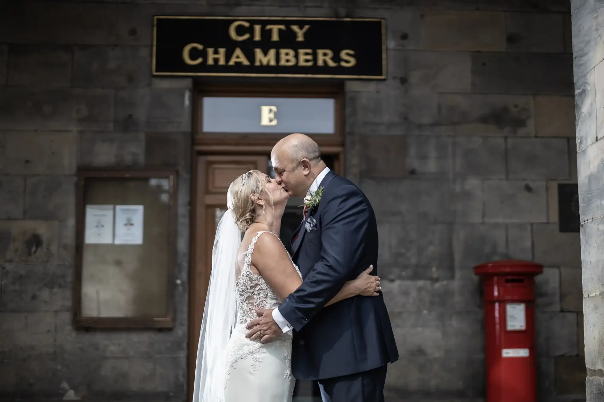 A bride and groom kiss in front of a building labeled "City Chambers," with a red post box nearby.