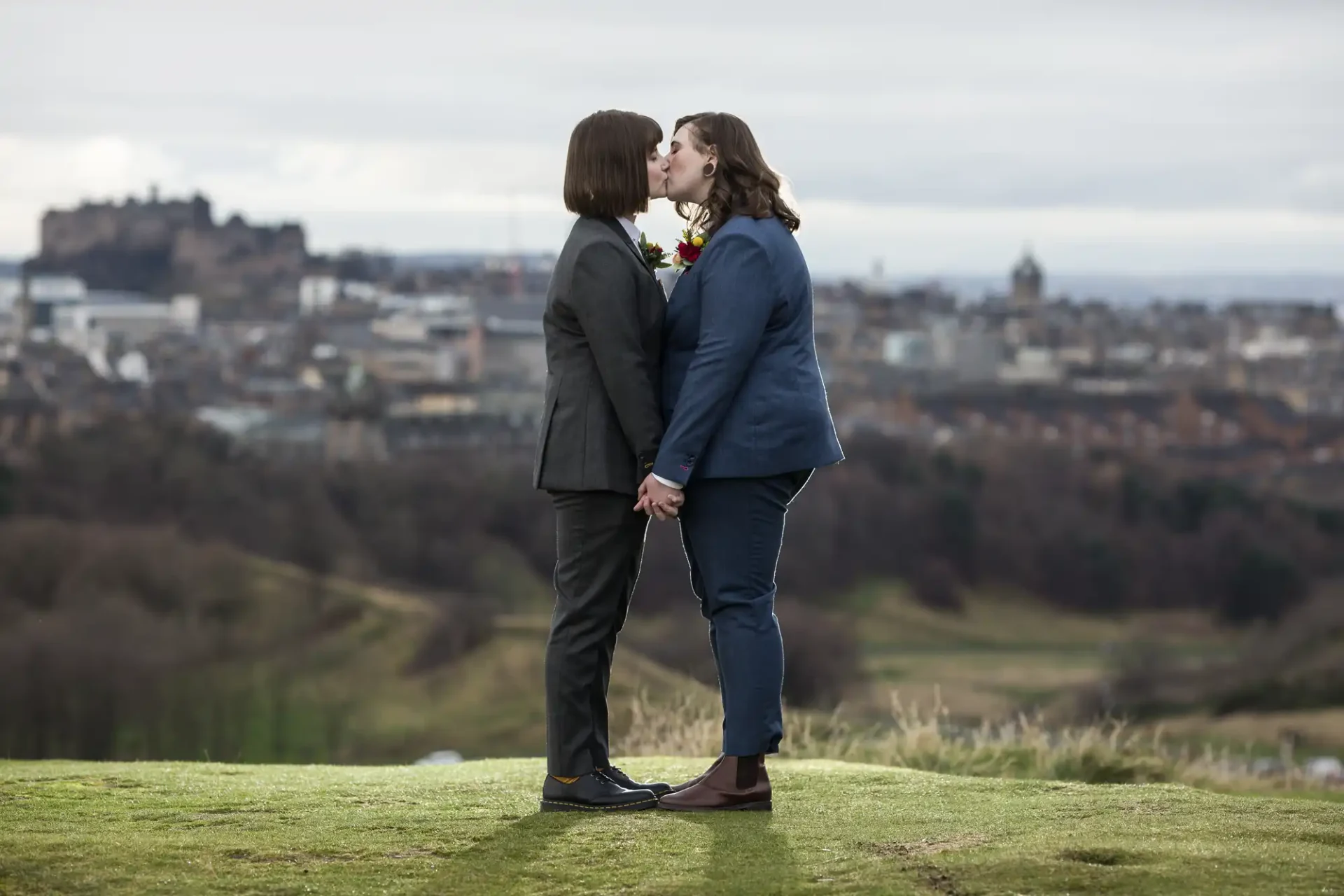 Two people in suits share a kiss while standing on grass with a cityscape in the background.