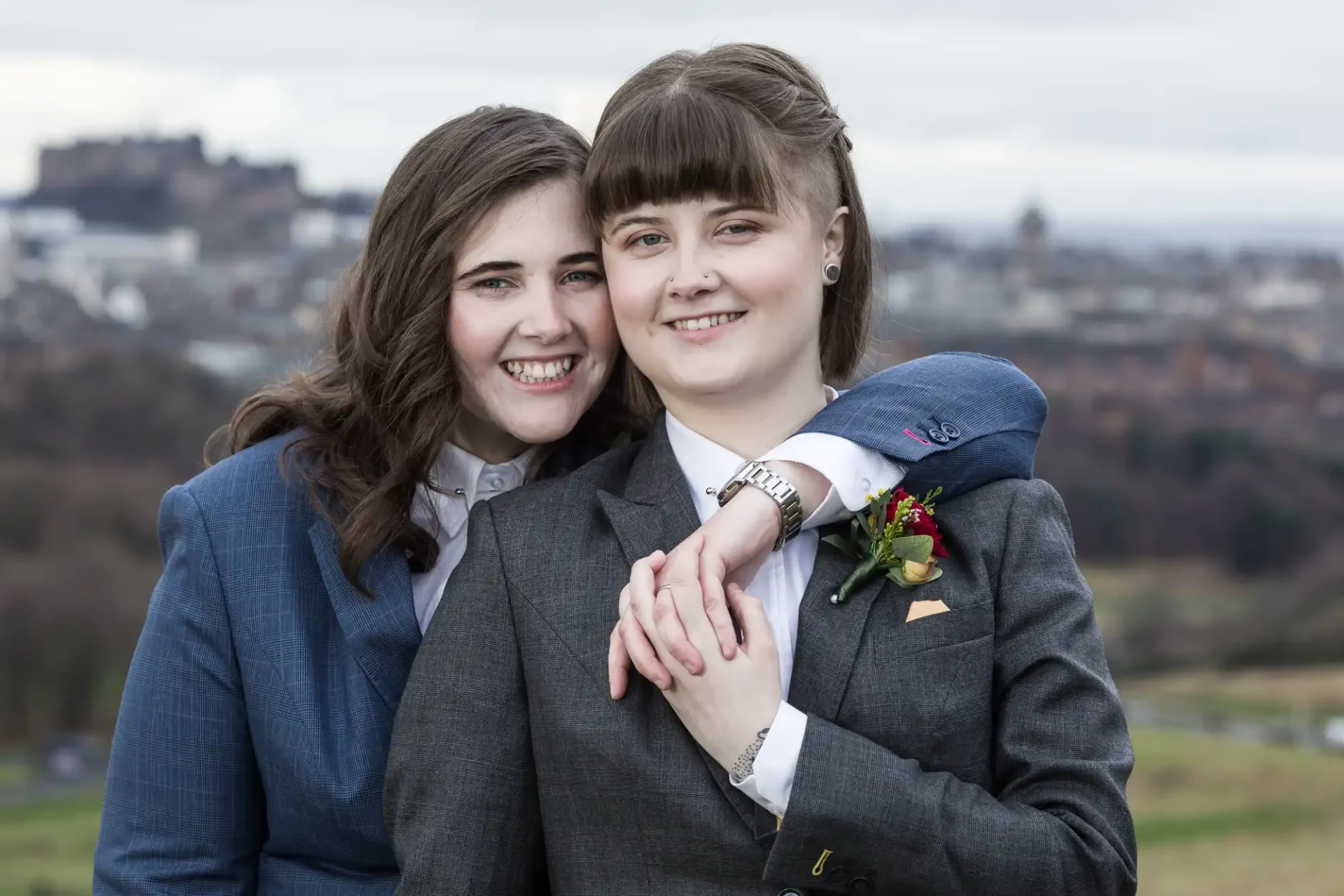 Two people stand closely, smiling, in formal attire. A castle is visible in the background under a cloudy sky.