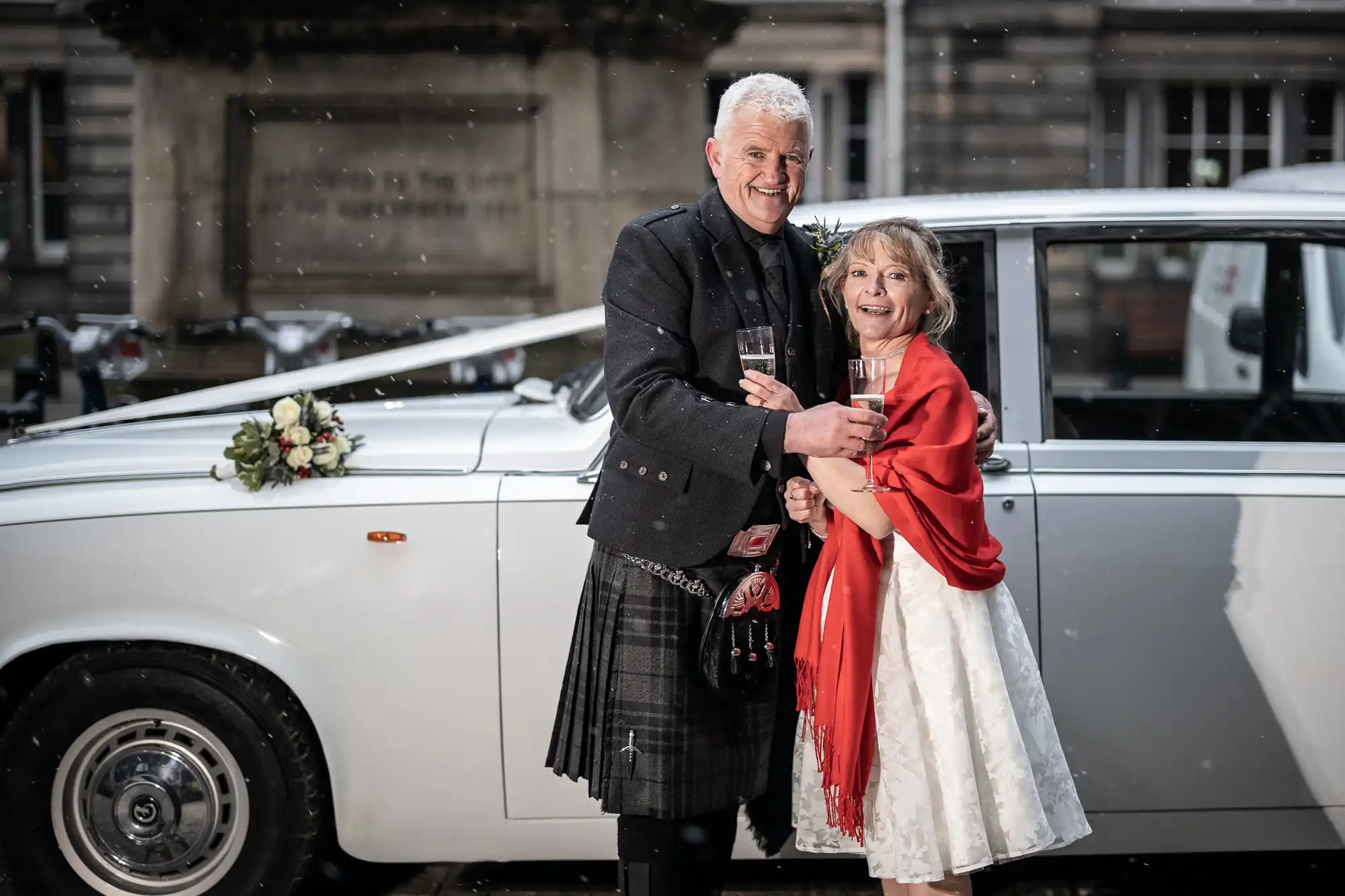 A couple stands in front of a white car decorated with flowers, holding champagne glasses and smiling, with light snow falling around them.