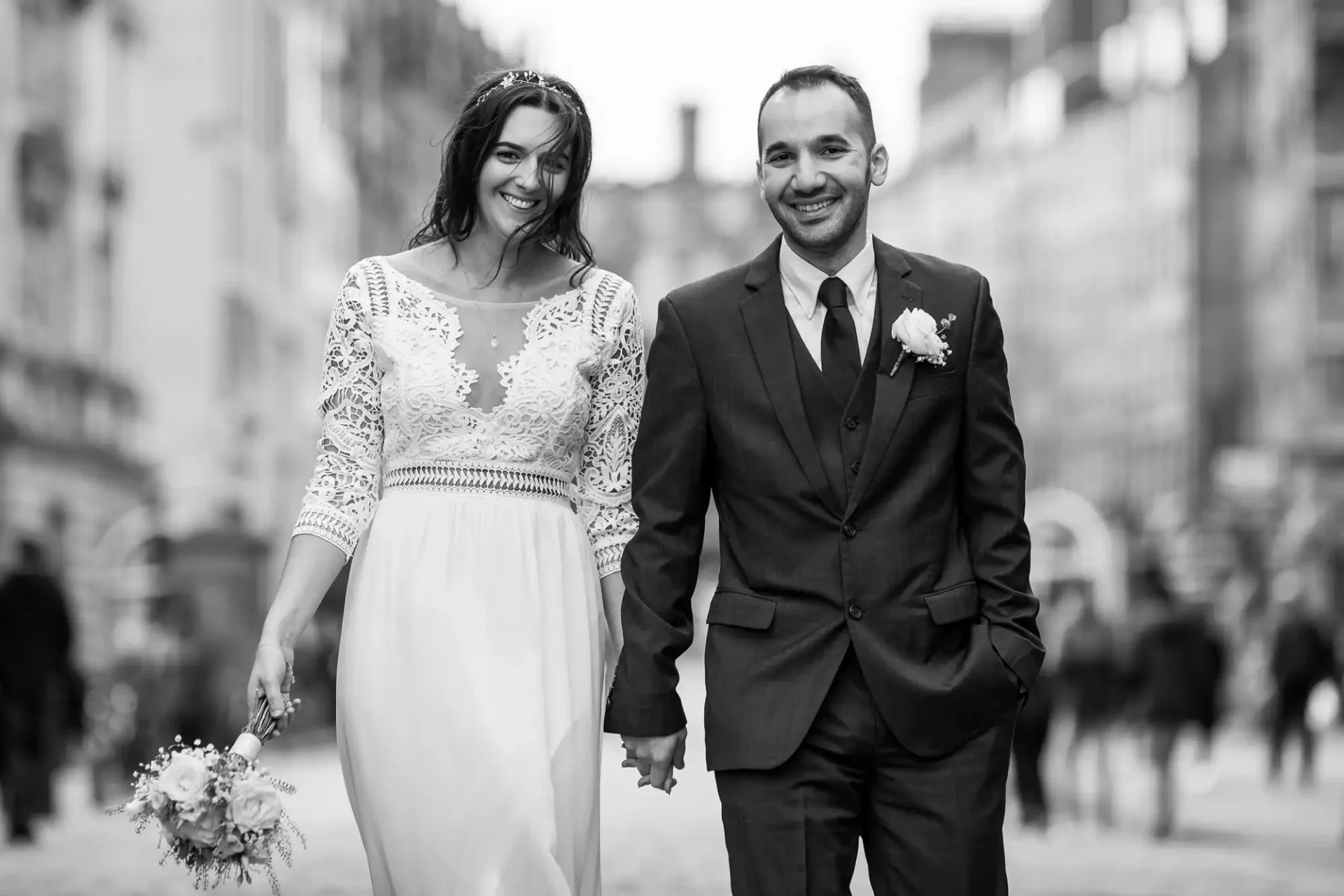 A couple in wedding attire walks hand in hand on a city street, smiling. The woman holds a bouquet, and the background shows blurred buildings and people.