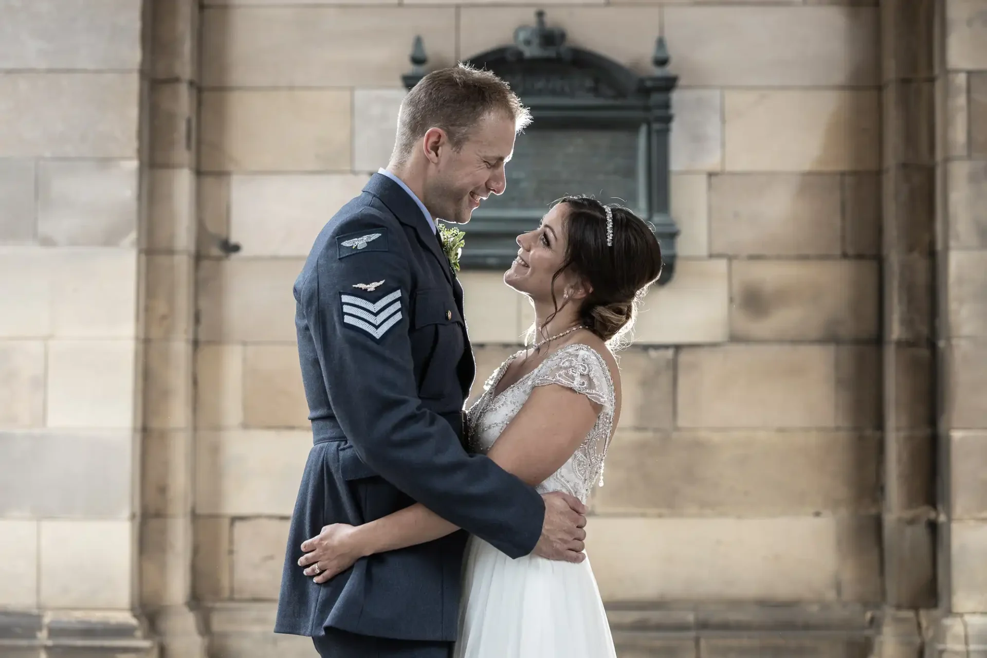 A couple in wedding attire, the groom in a military uniform and the bride in a white dress, stand facing each other in front of a stone wall.