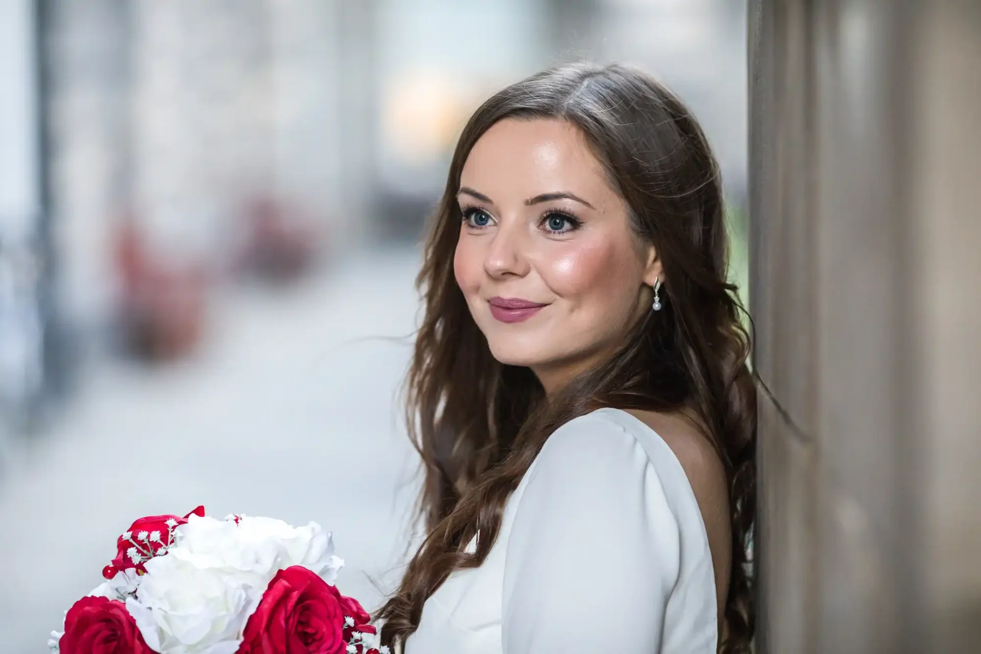 A woman in a white dress holds a bouquet of red and white flowers while leaning against a wall, gazing to her left.