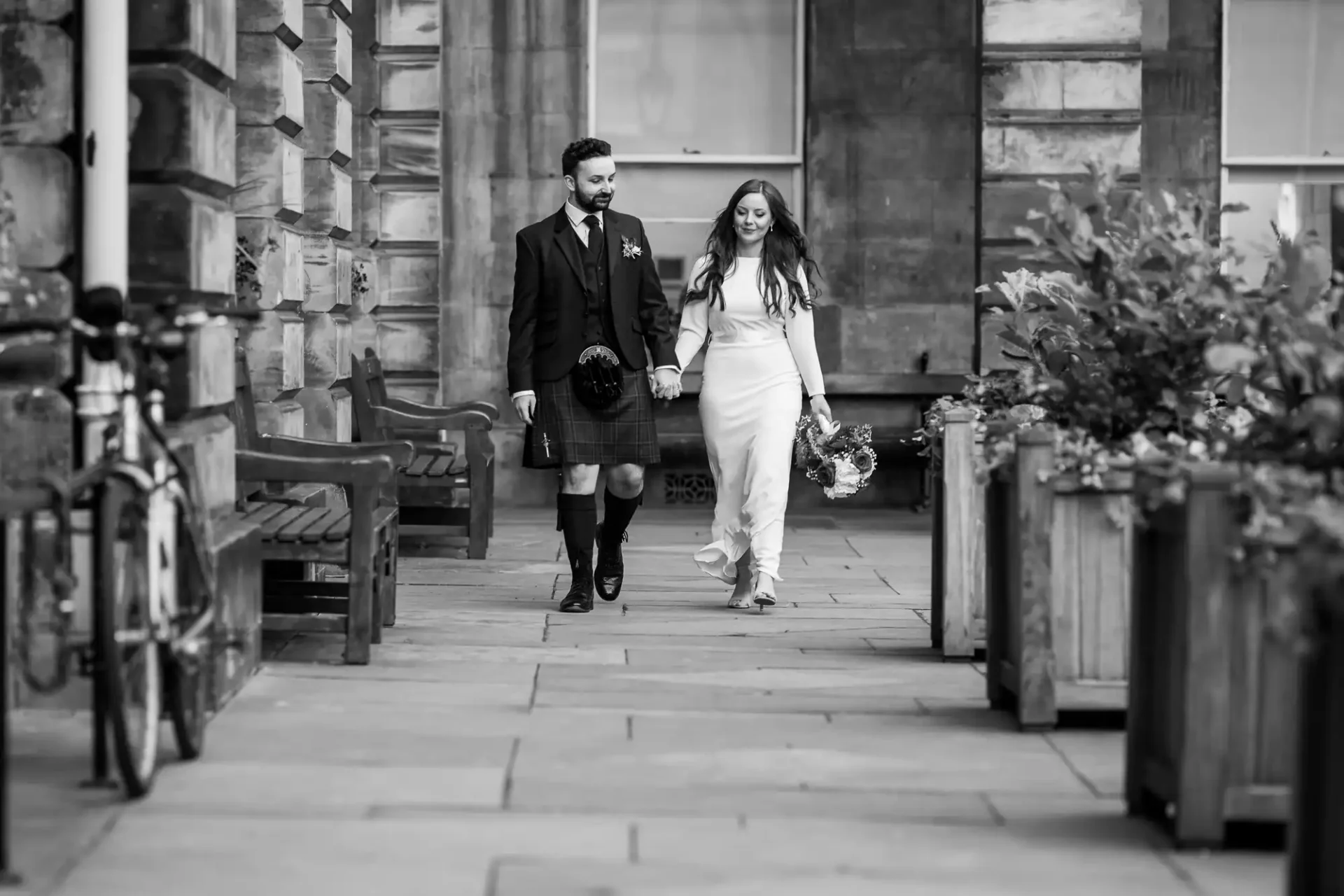 A couple walks hand in hand along a stone pathway lined with benches and plants. The man wears a kilt, and the woman holds a bouquet. Black and white photo.