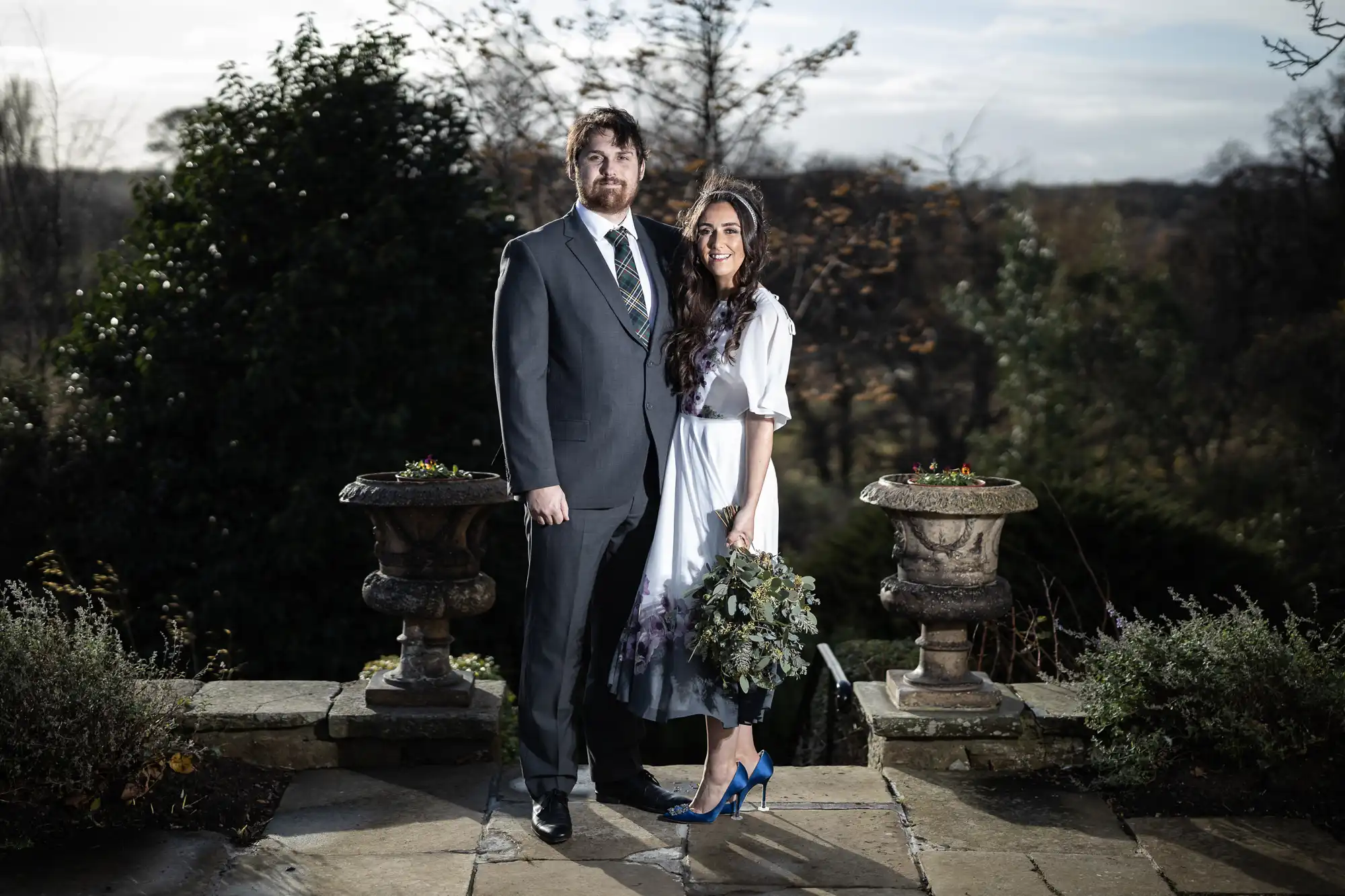 A man in a grey suit and a woman in a white dress with blue heels stand close together, smiling, on a stone patio surrounded by greenery. The woman holds a bouquet; two decorative stone urns flank the pair. Trees and overcast sky fill the background.