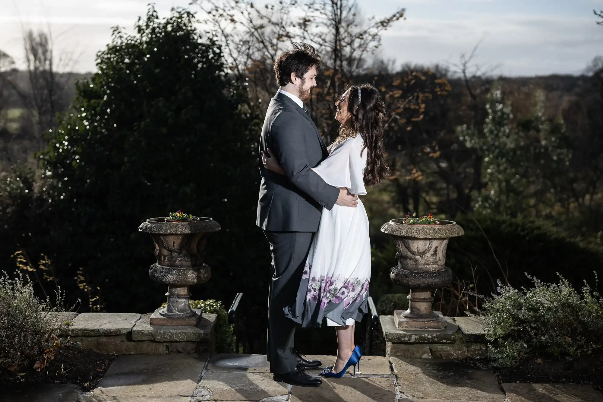 A man in a dark suit and a woman in a white dress with floral patterns and blue heels stand facing each other, smiling and holding each other’s waists, outdoors on stone steps between two planters, with trees and greenery in the background.