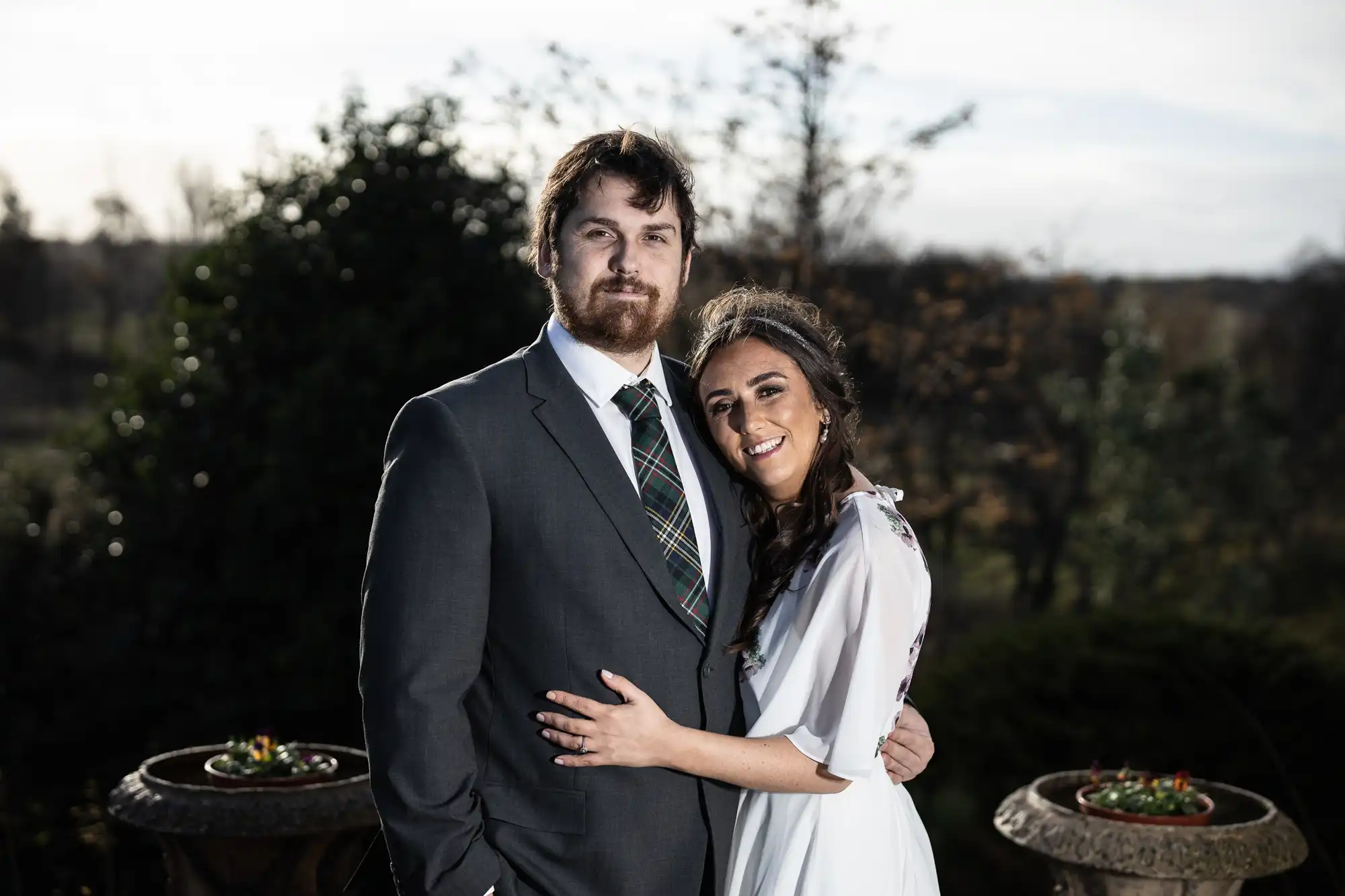 A man in a dark suit with a checked tie stands beside a woman in a white dress. They smile and pose outdoors with his arm round her. The background features blurred greenery and trees, with overcast sky and two stone planters on either side.