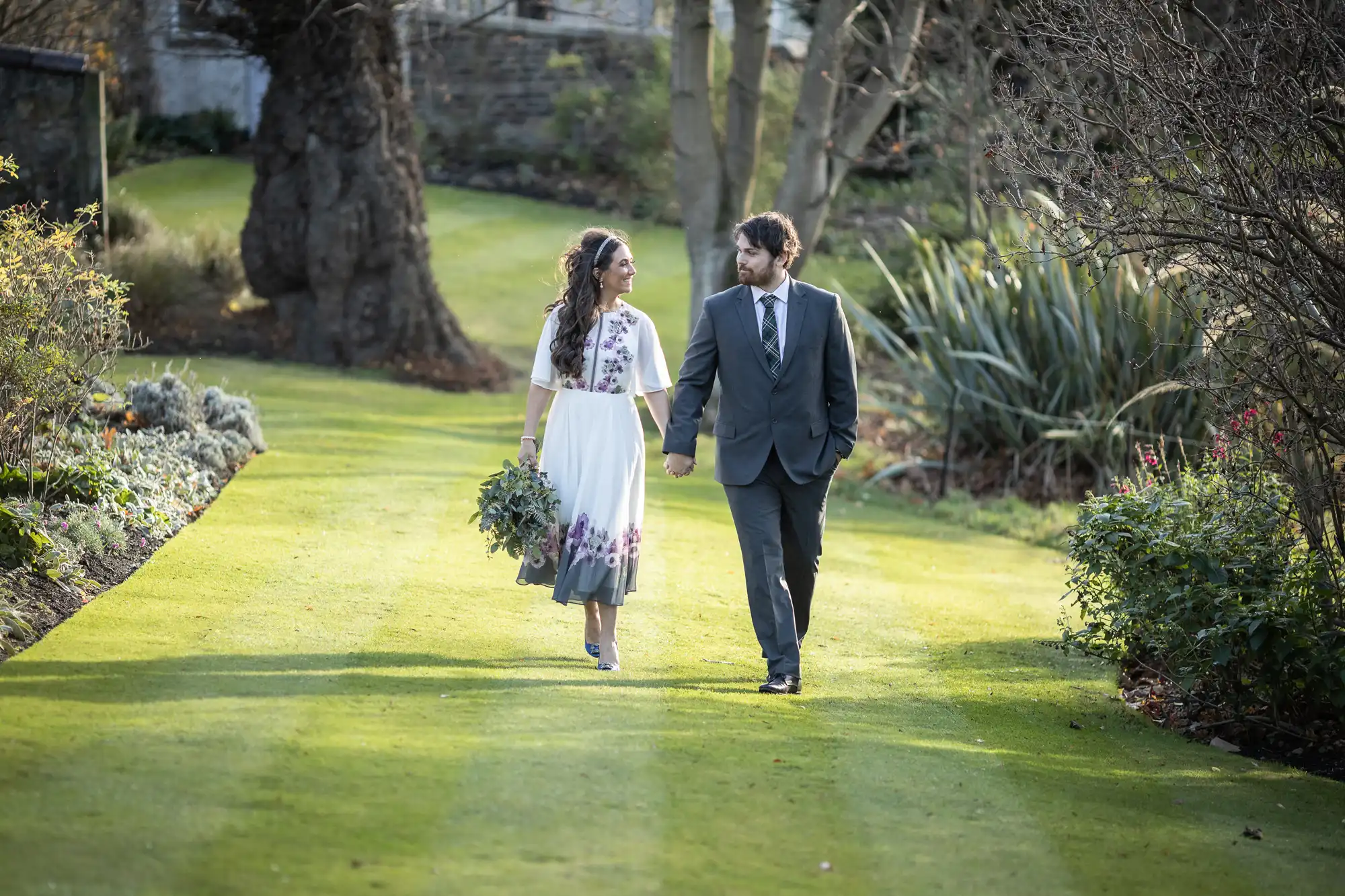 A couple walk hand in hand on a sunlit garden path. The woman wears a white dress with purple flowers and holds a bouquet. The man wears a grey suit. Green grass and trees surround them, with shrubs and flowers lining the path and sunlight filtering through branches.